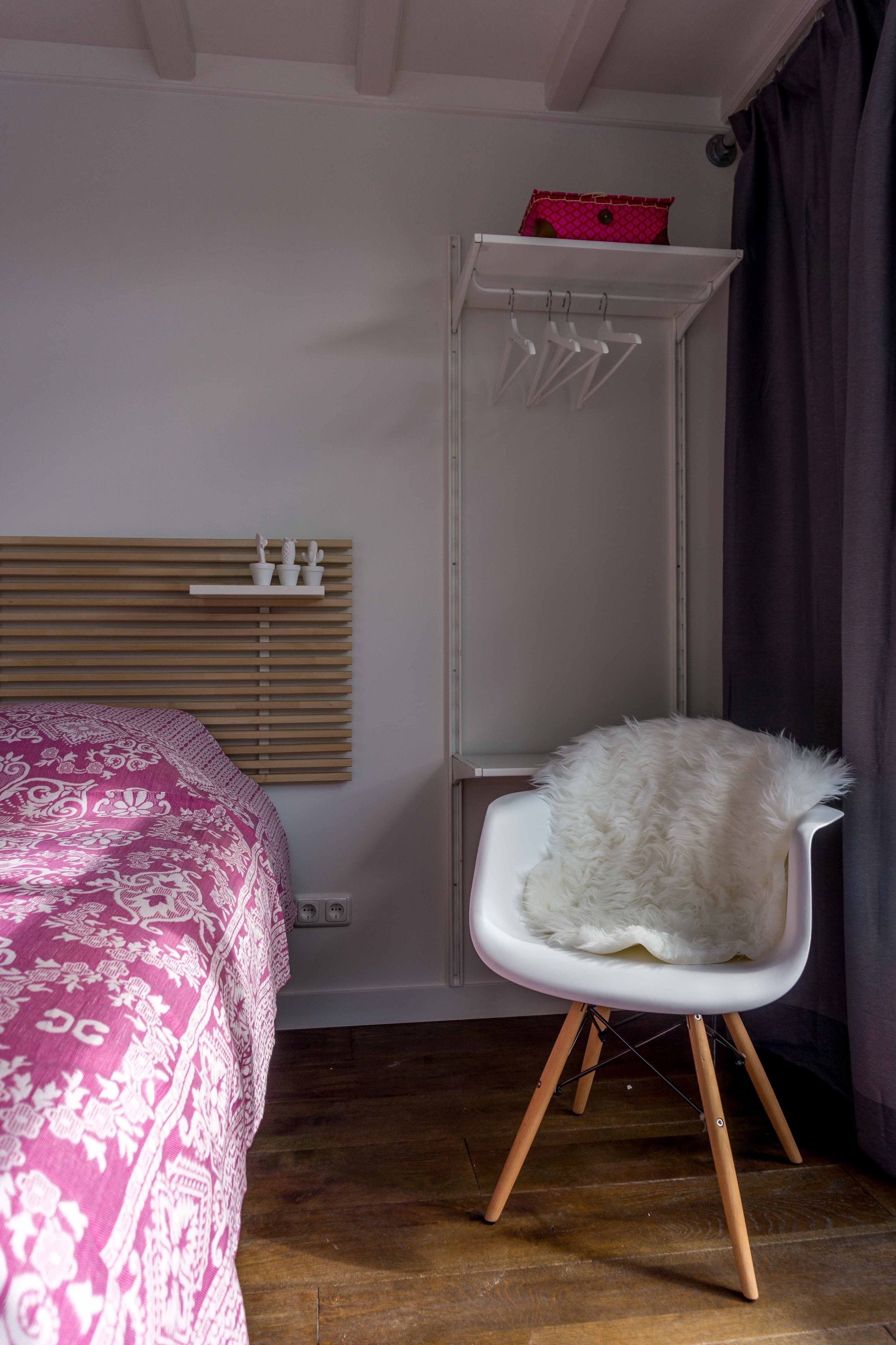 A bedroom with a bed, a white chair with a fluffy seat, a white wardrobe with a pink item on top, and a window with blinds.