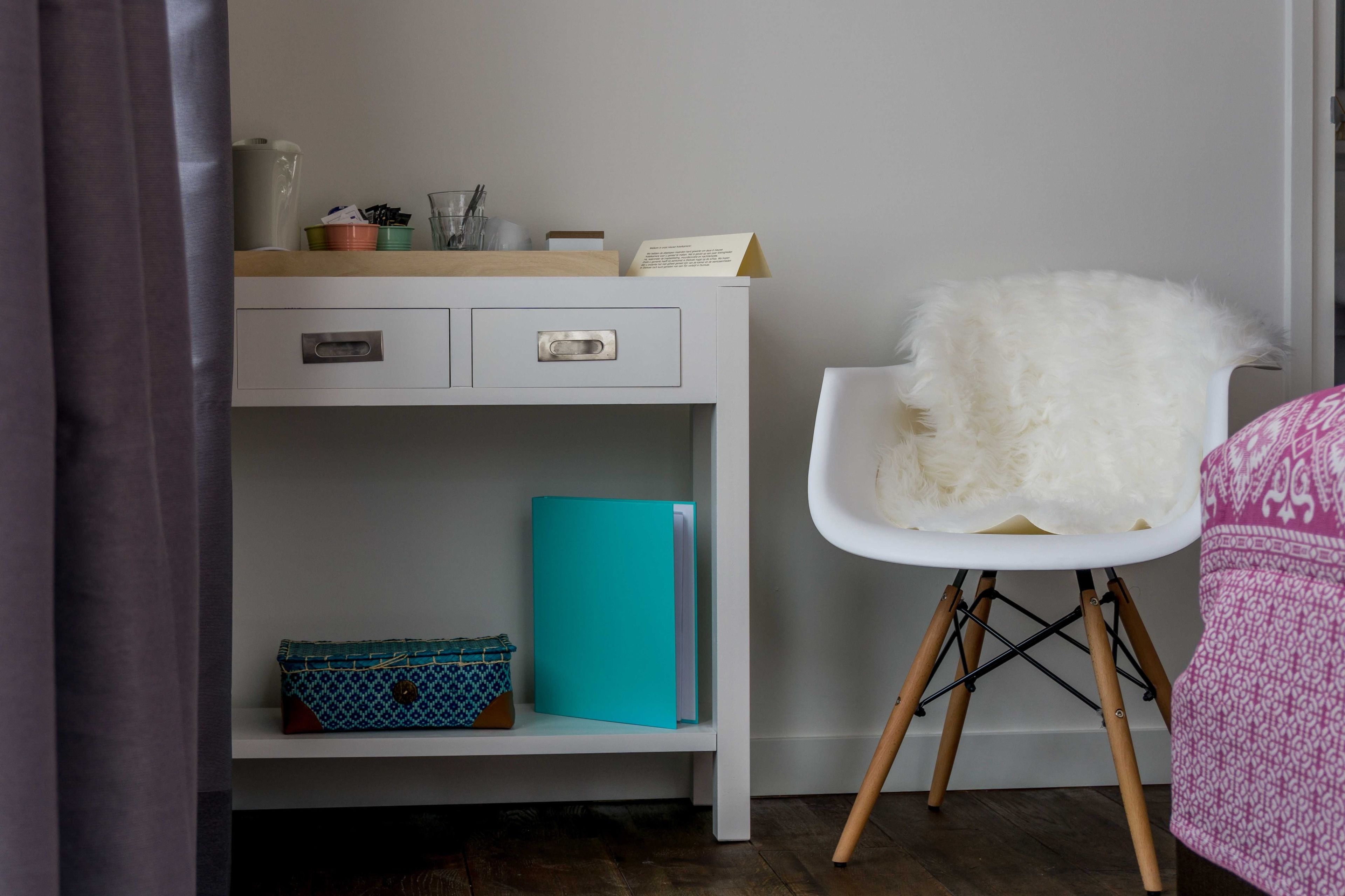 Un escritorio blanco con cajones, una carpeta azul verdosa y una silla blanca con patas de madera.