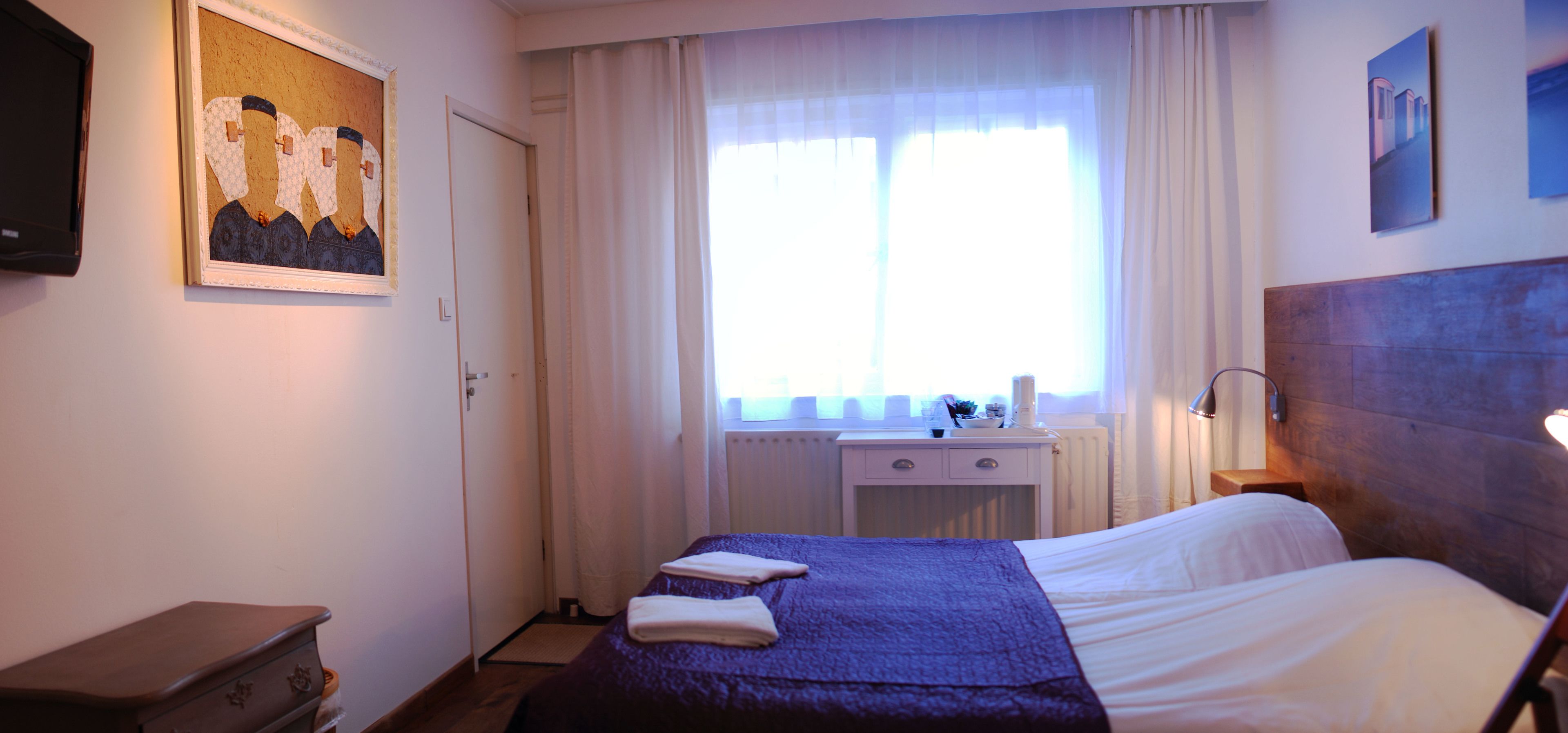 Une chambre d’hôtel avec un lit, une table de chevet, une fenêtre avec des rideaux, un tableau au mur et une tête de lit en bois.