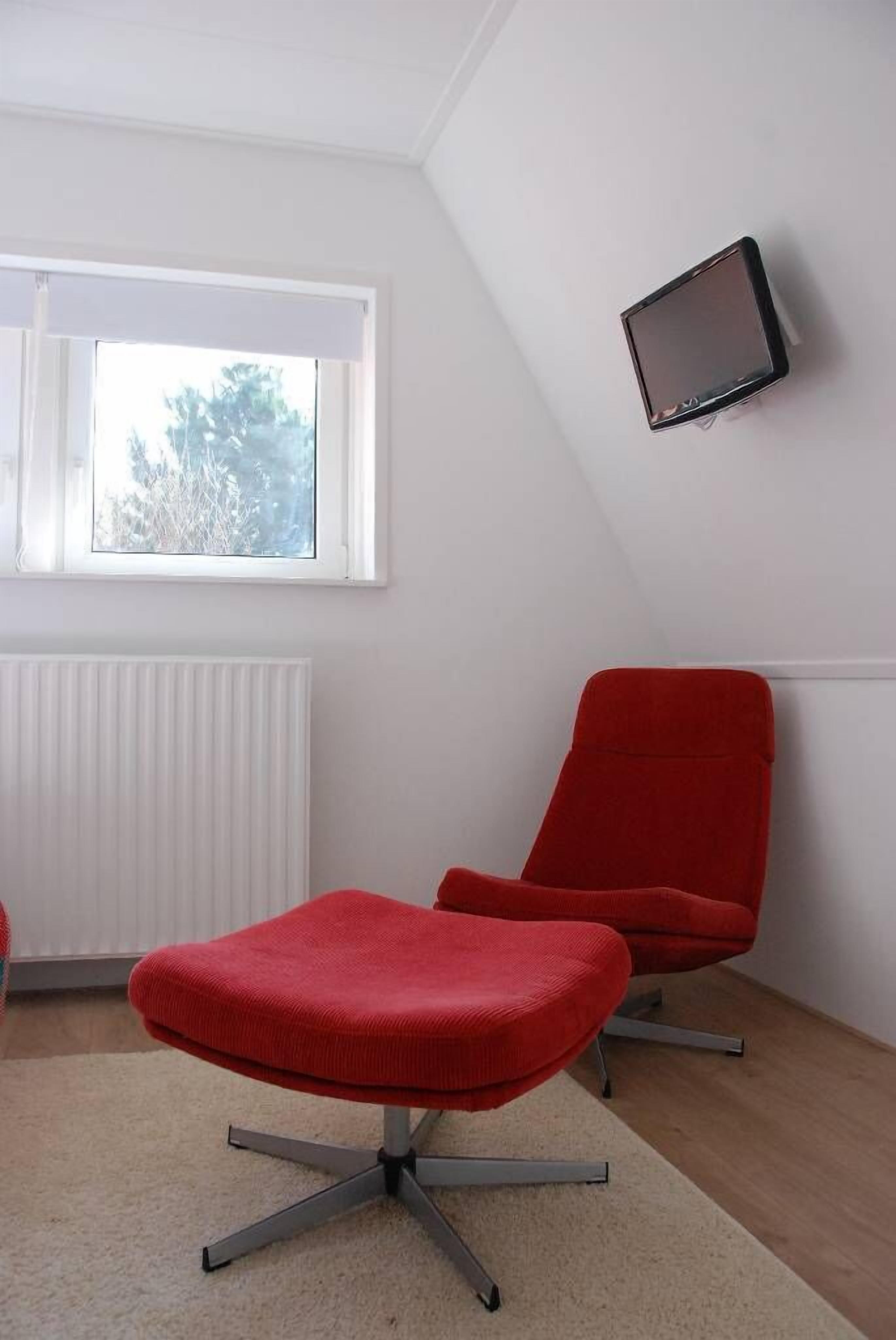 A room with a red chair, a red ottoman, a white radiator, and a TV mounted on the wall.