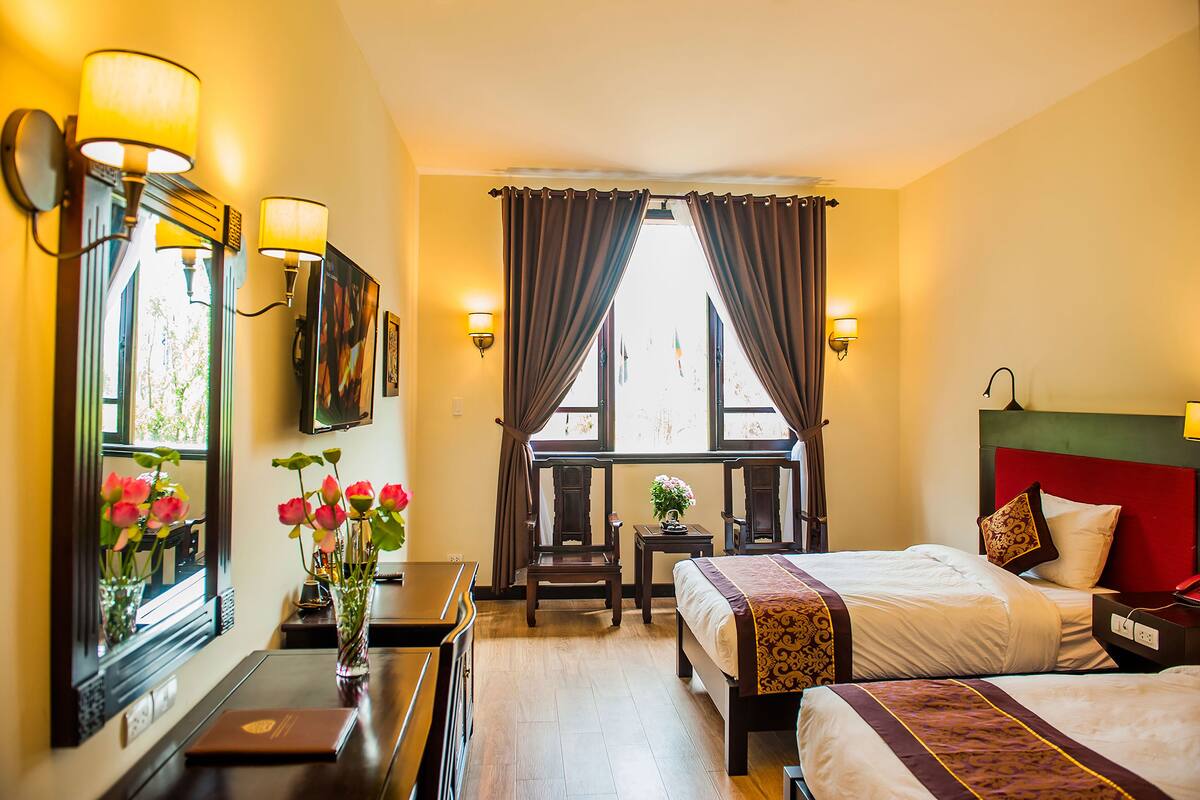 Une chambre d’hôtel avec deux lits, un bureau, une chaise, une petite table ornée d’un vase de fleurs et une fenêtre avec des rideaux.