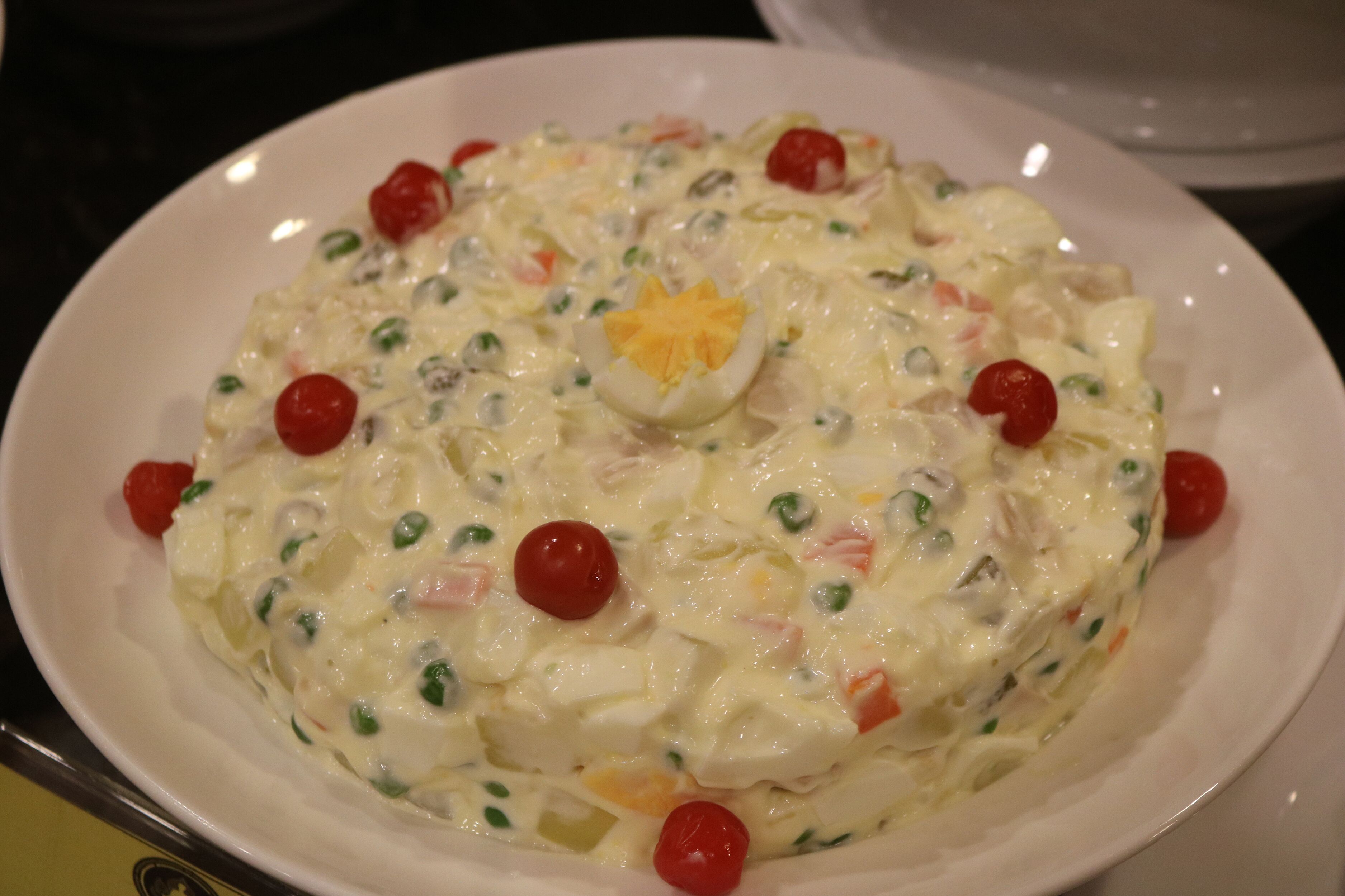 A large white plate of creamy potato salad with boiled eggs, cherry tomatoes, and green peas.