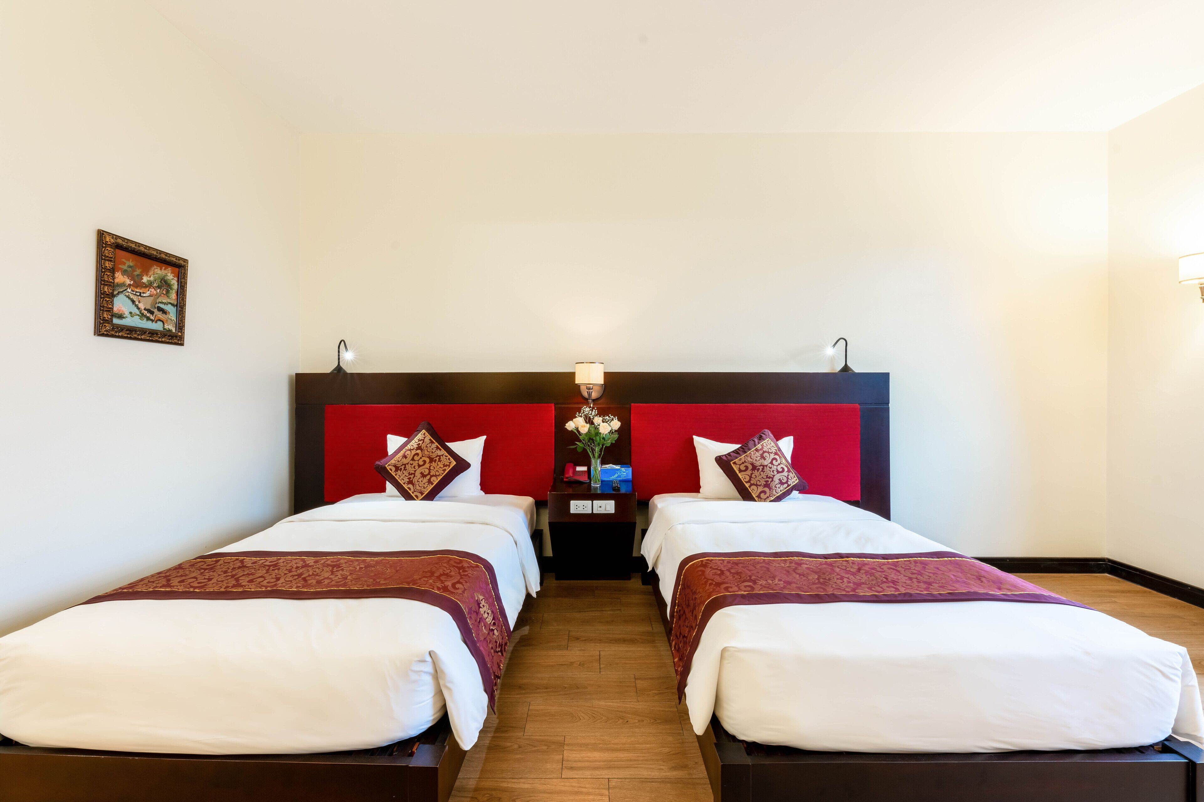 Habitación de hotel con dos camas, cabecero rojo, ropa de cama blanca, una mesita de noche con un jarrón de flores y suelo de madera.