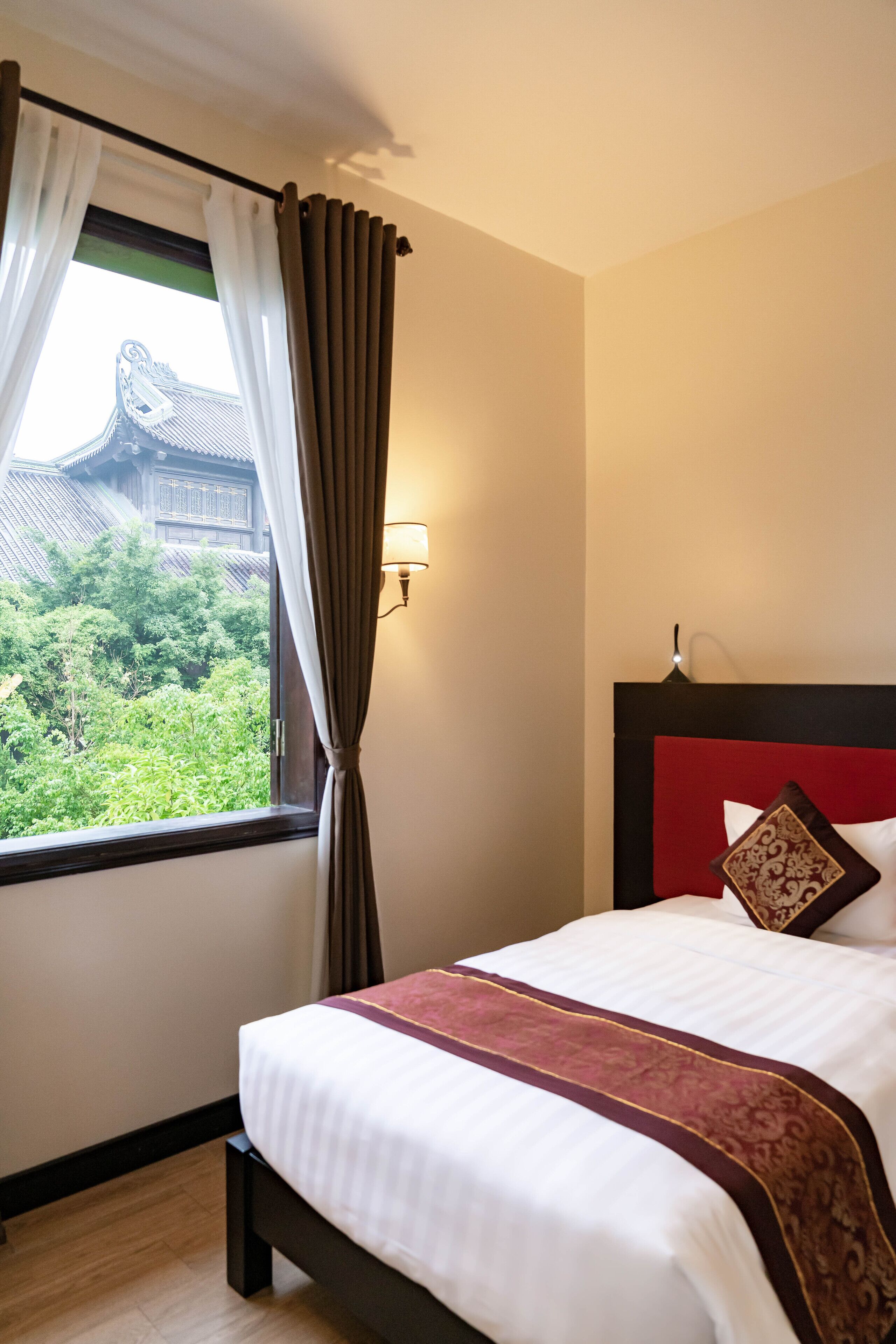 A hotel room with a bed, a window with curtains, a wall-mounted lamp, and a view of a traditional building and greenery outside.