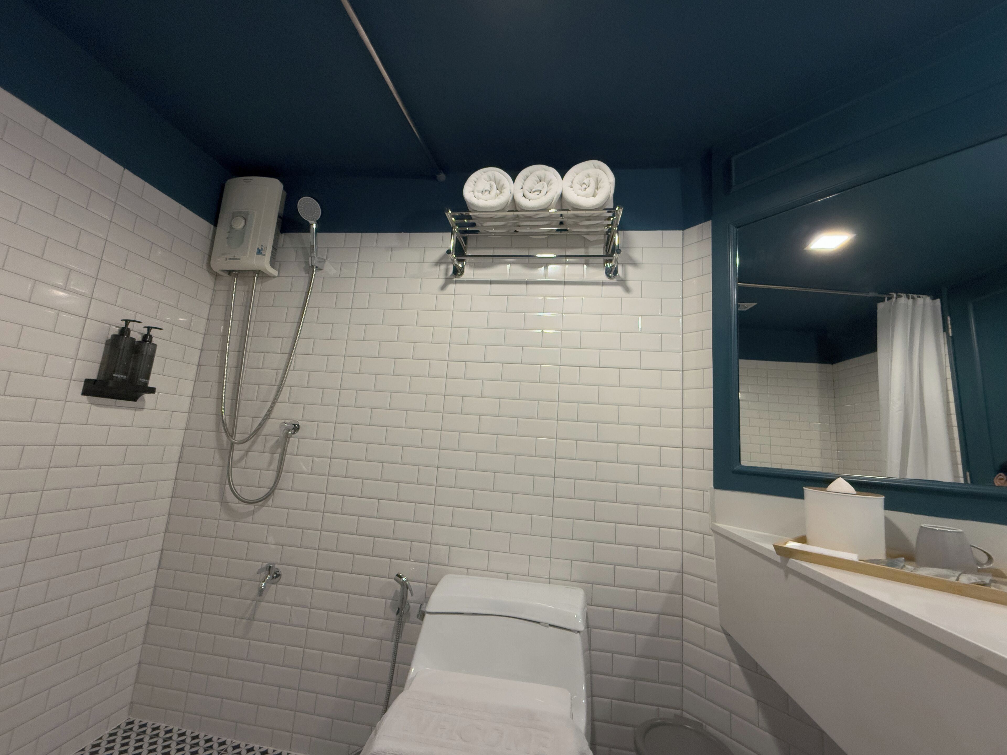 A bathroom with white tiled walls, a shower, a toilet, and a sink with a mirror above it.