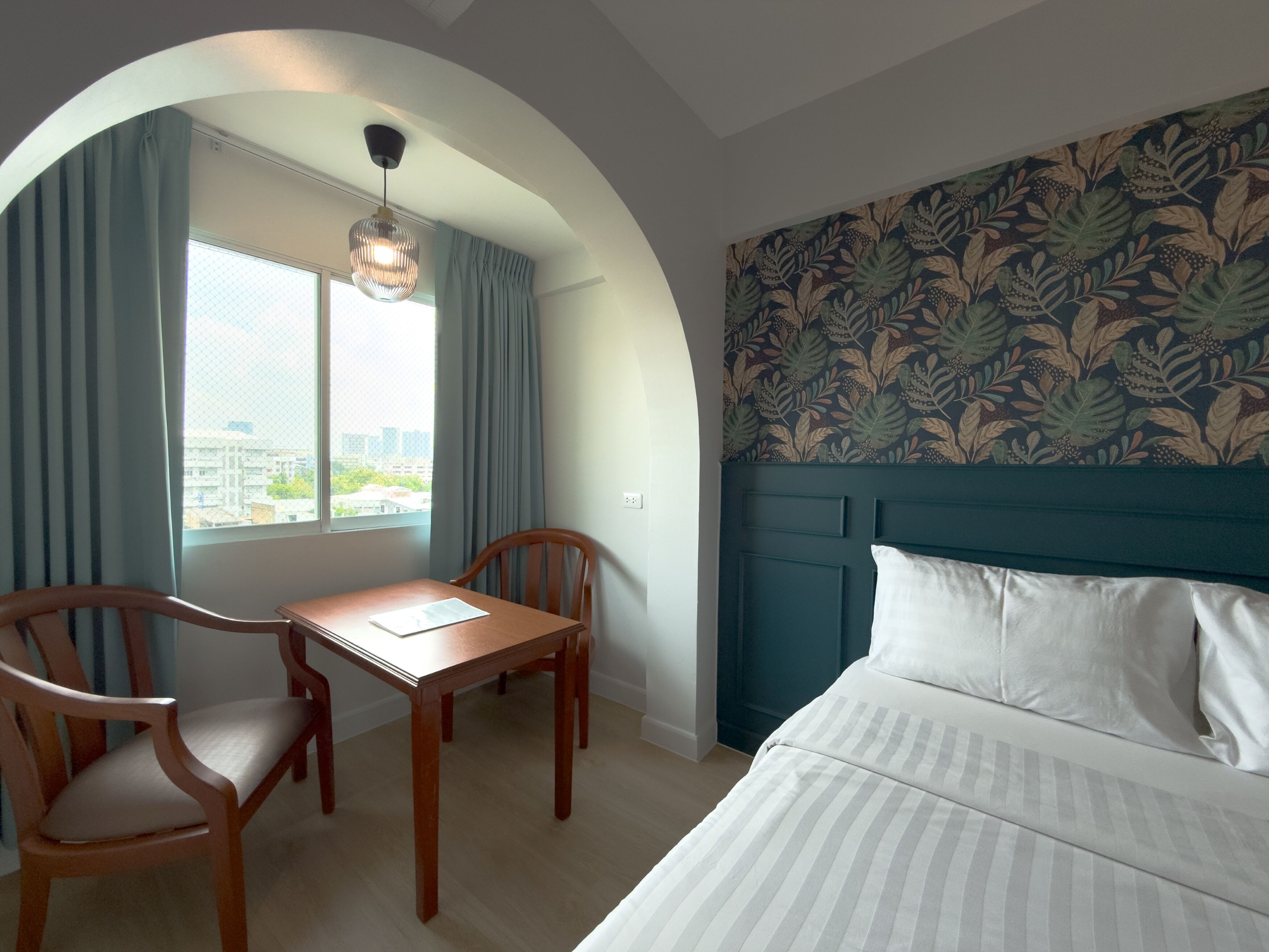 A hotel room with a bed, a table, two chairs, a window with curtains, and a patterned wall.