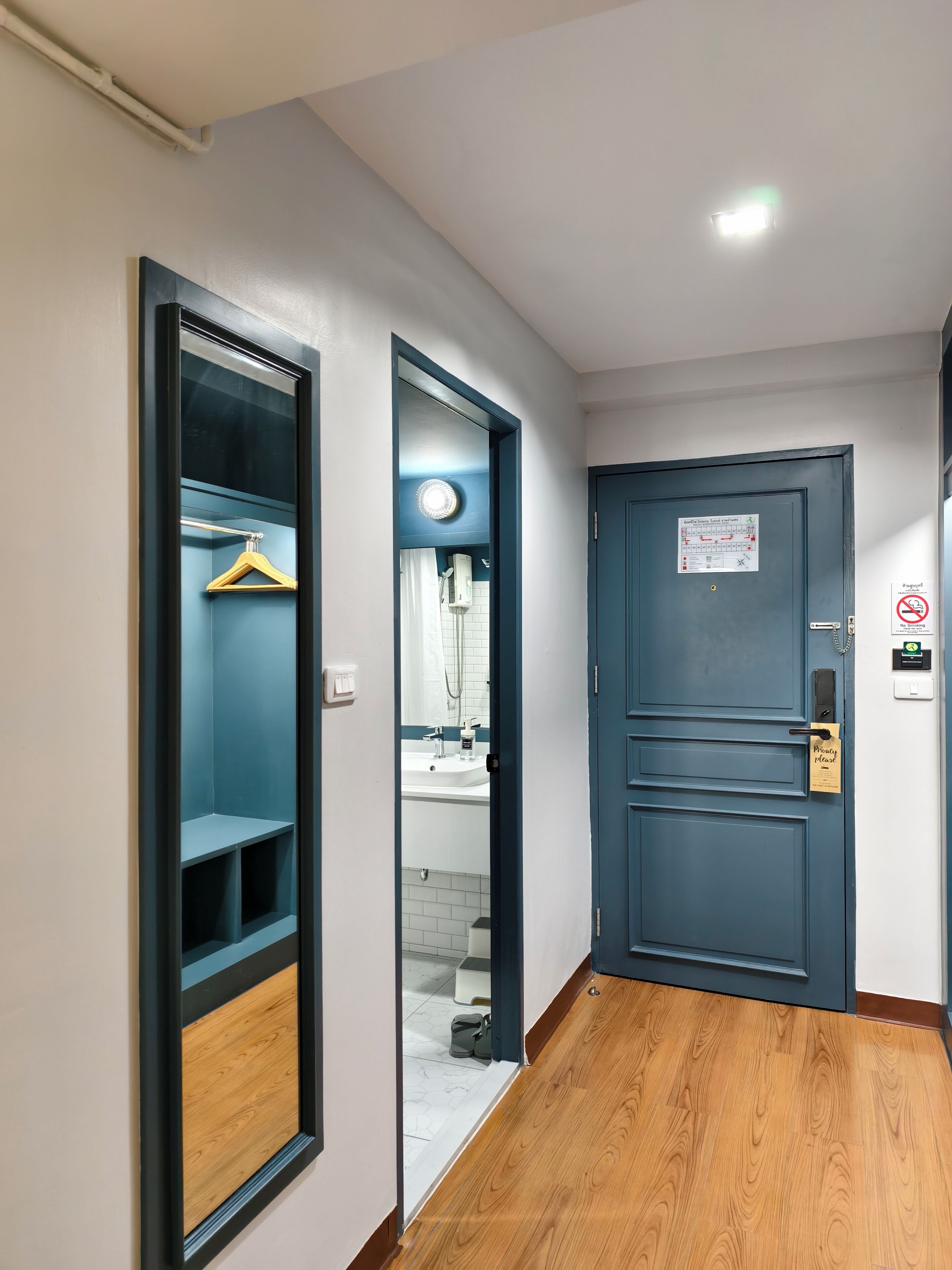 A hallway with a wooden floor, a blue door with a 'Please Wait' sign, and a mirror reflecting a bathroom with a sink and shower.
