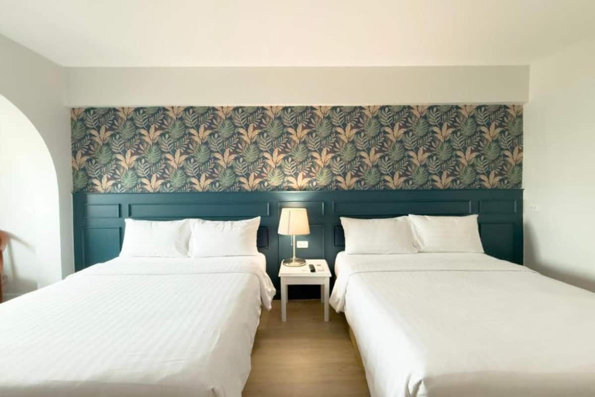 A hotel room with two beds, a nightstand with a lamp, and a patterned wallpaper.