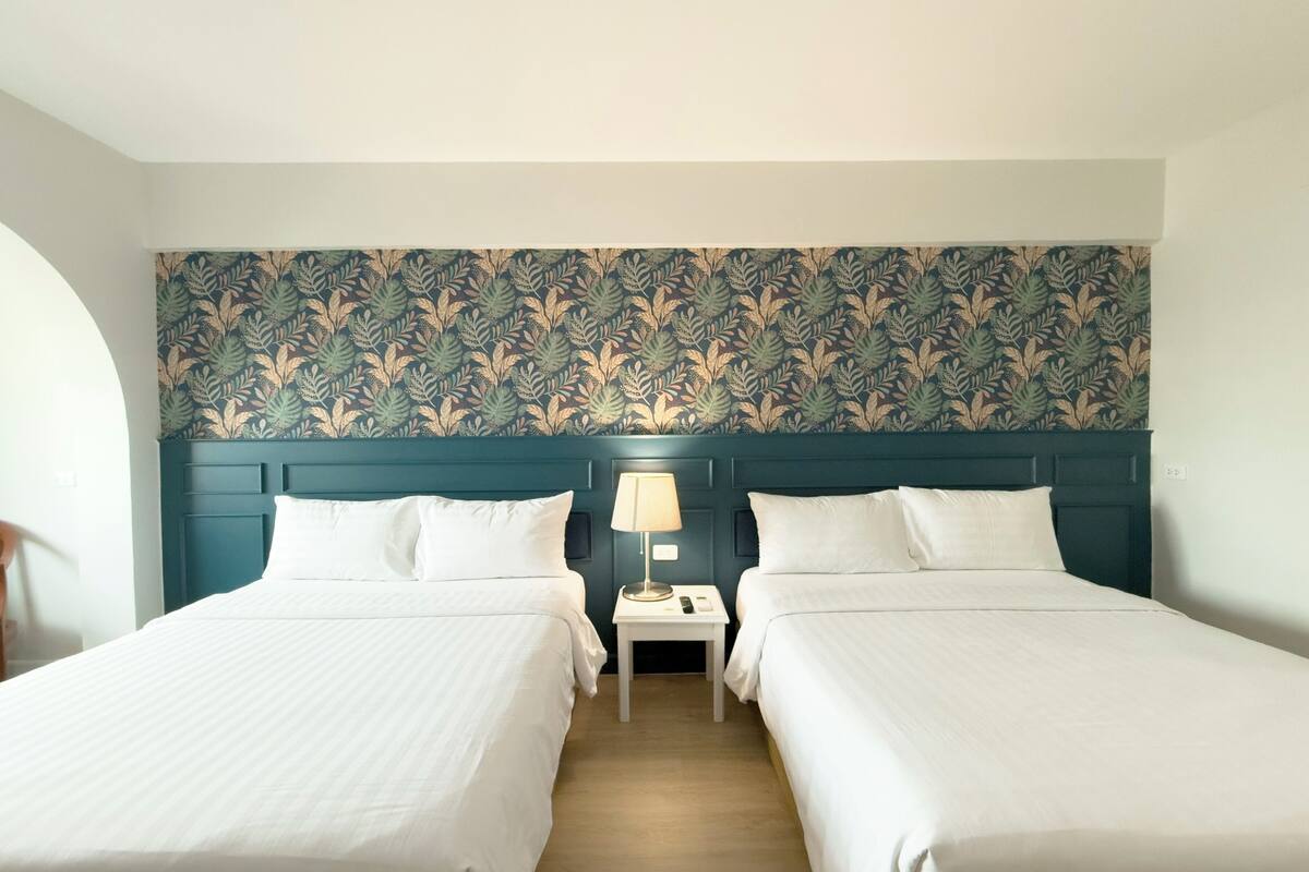 A hotel room with two beds, a nightstand with a lamp, and a floral-patterned headboard.