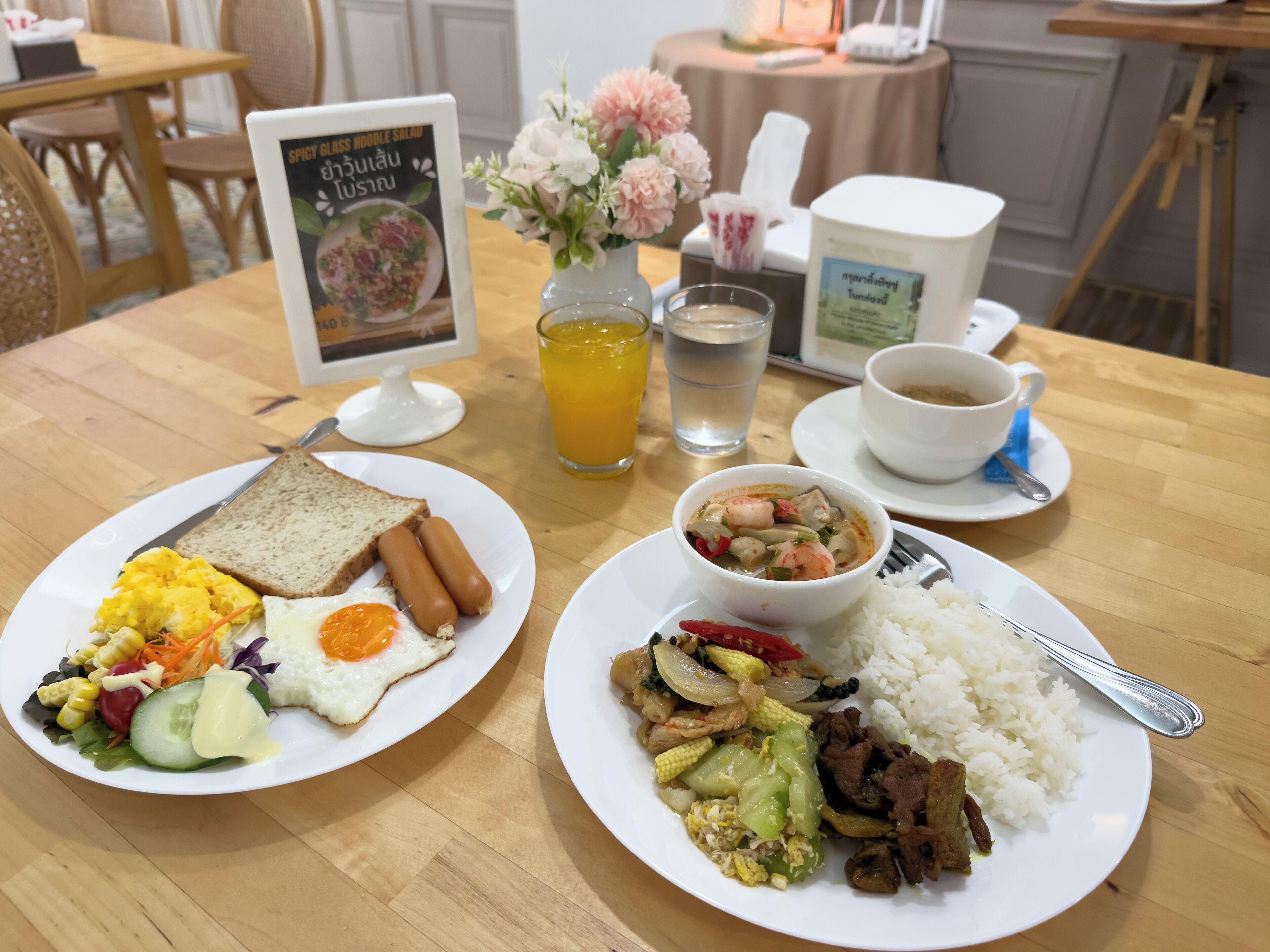 Una mesa de desayuno con dos platos de comida, un menú, un jarrón de flores, un vaso de jugo de naranja, una taza de café y un vaso de agua.