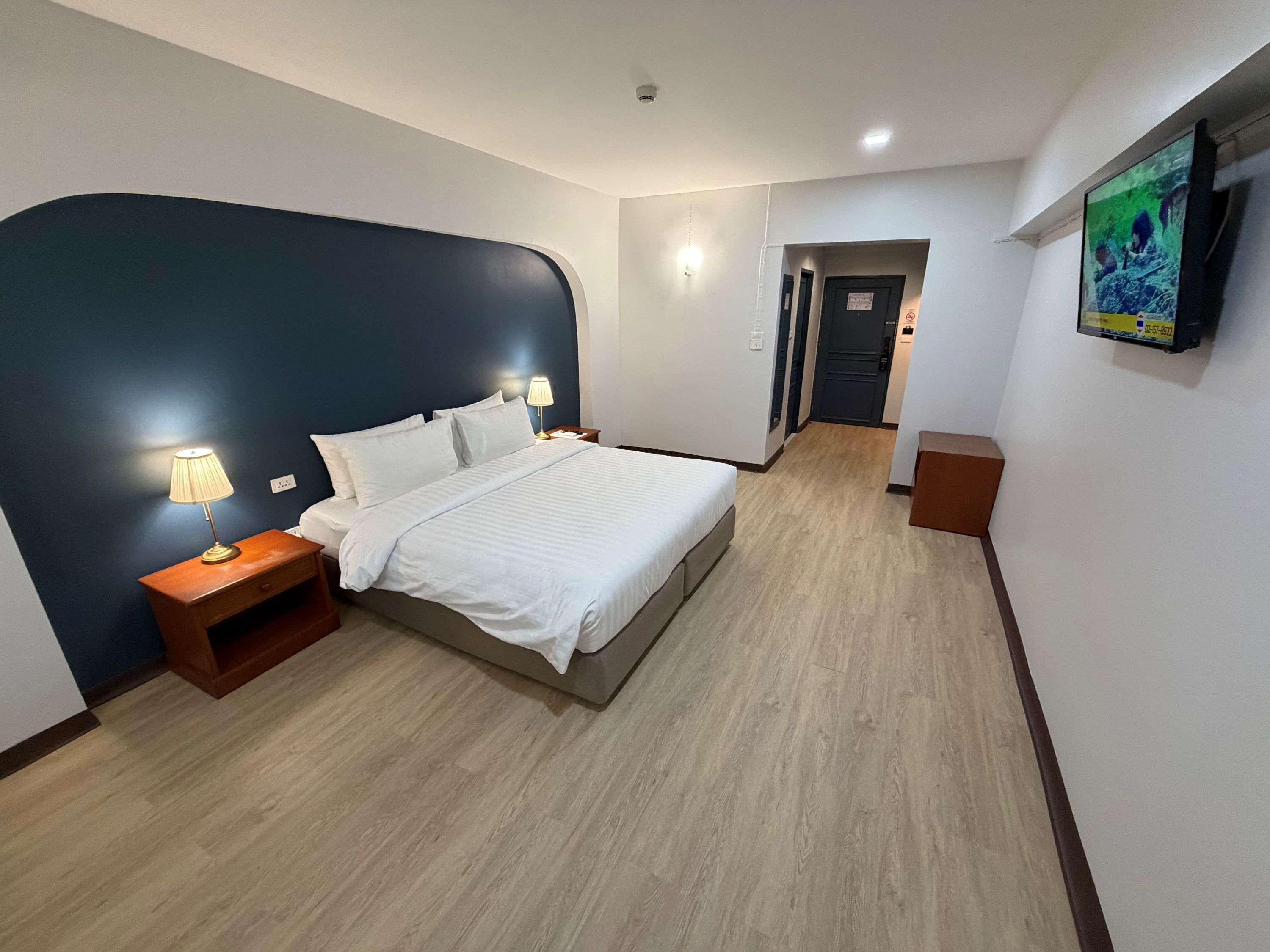 A hotel room with a bed, bedside lamps, a wooden nightstand, a flat-screen TV mounted on the wall, and a wooden floor.