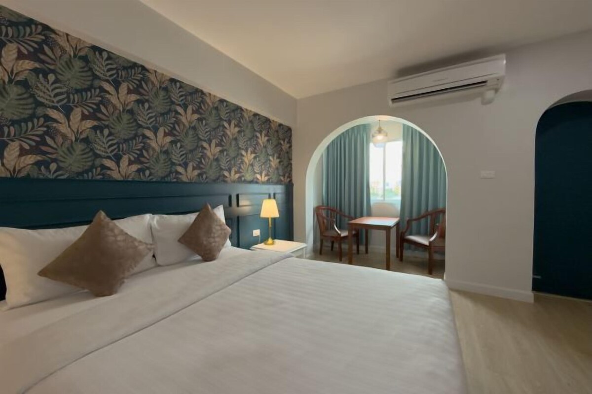 A hotel room with a bed, a desk, chairs, a lamp, and a view through an arched window.