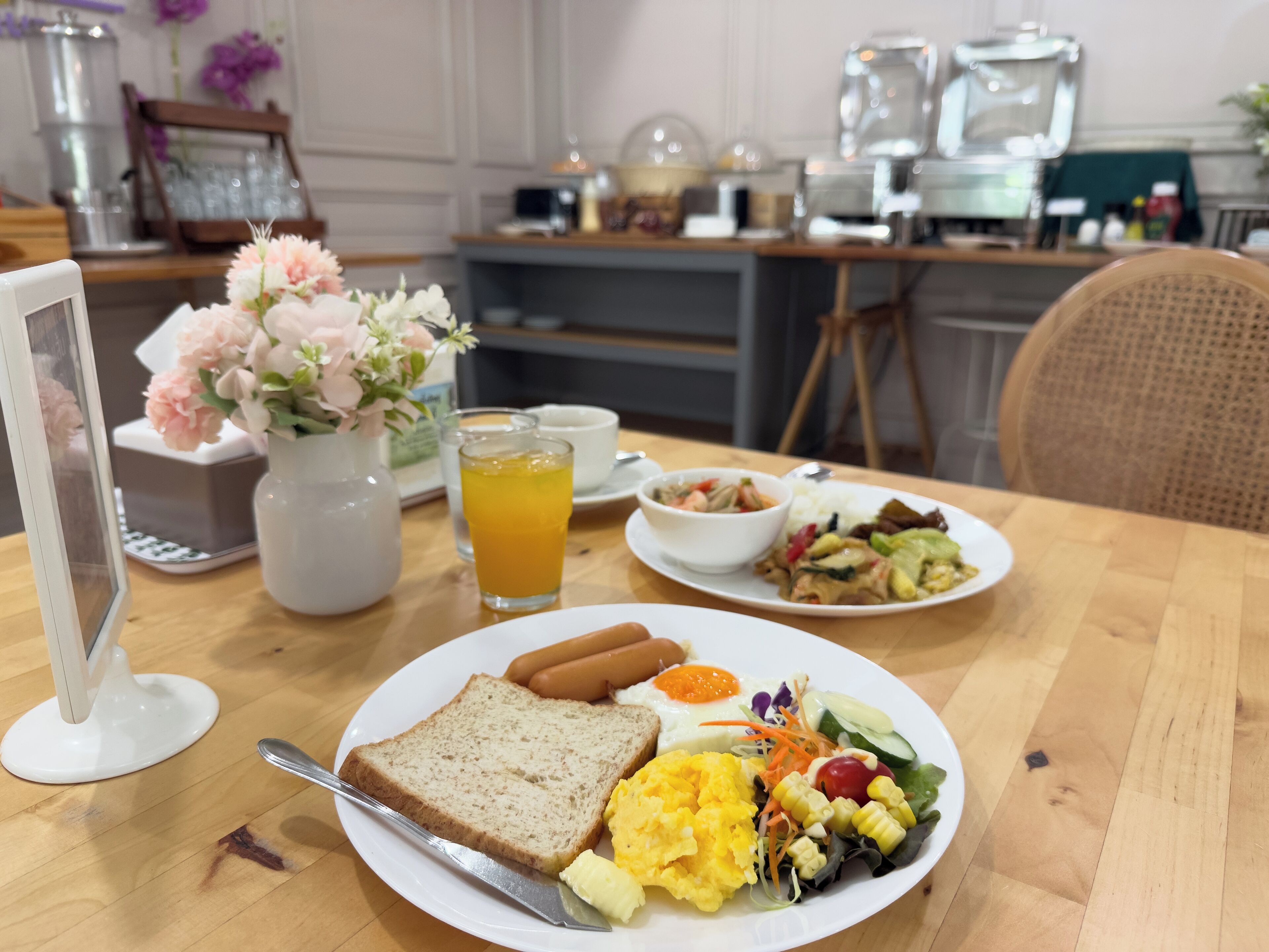 A breakfast table with a plate of toast, scrambled eggs, a salad, and a hot dog. There is a vase with flowers, a glass of orange juice, and a coffee maker in the background.