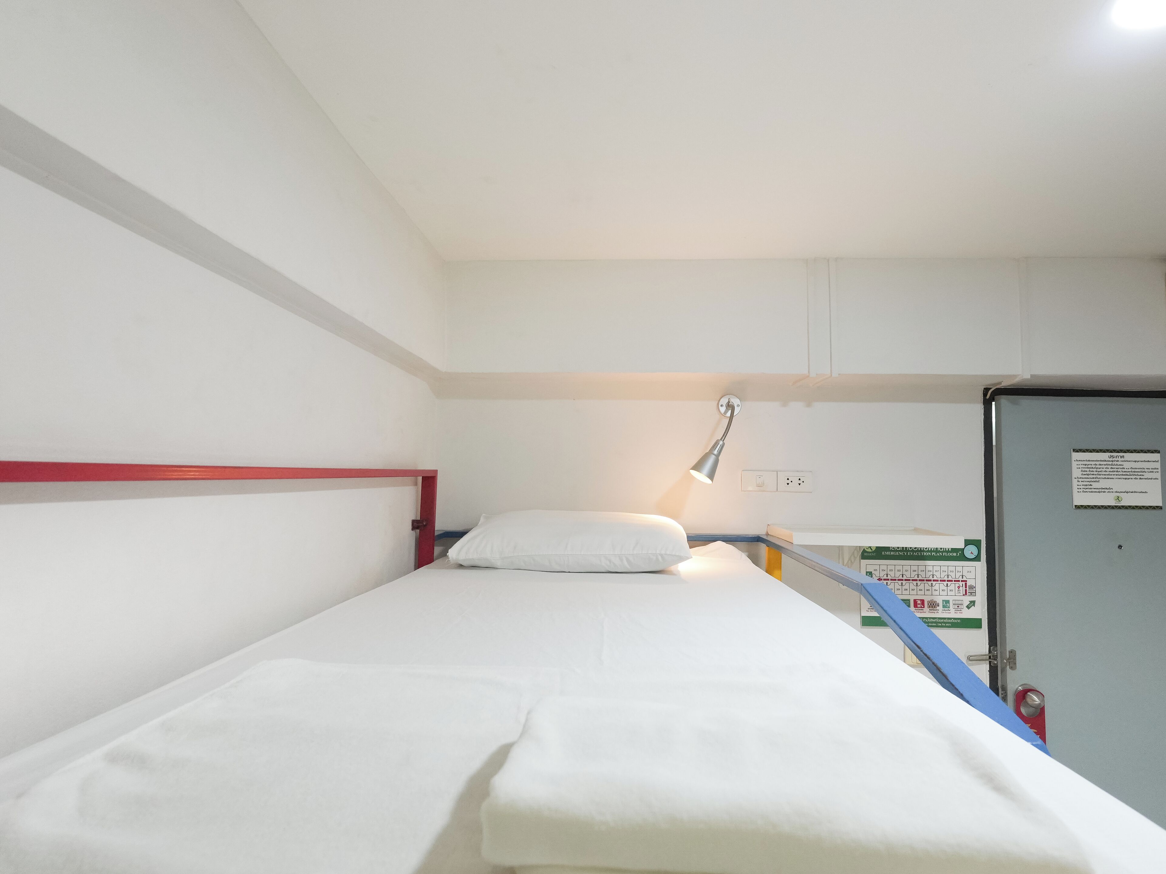 A single bed with a red headboard, a white pillow, and a bedside lamp. There is a small cabinet with a sign on it, and a blue railing is visible.
