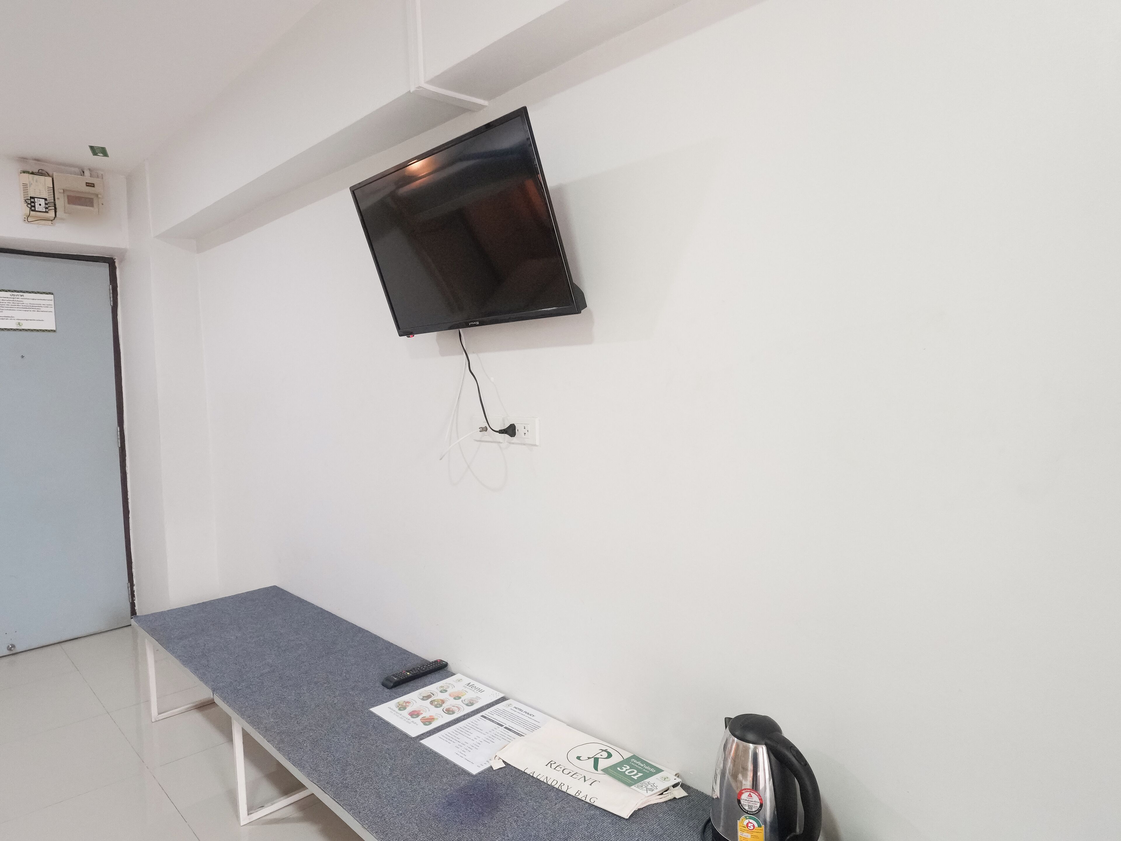 A room with a flat-screen TV mounted on the wall, a grey table with a remote control and a menu card, and a kettle on the table.