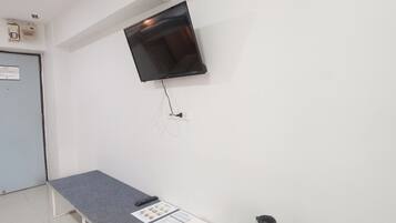 A room with a flat-screen TV mounted on the wall, a grey table with a remote control and a menu card, and a kettle on the table.