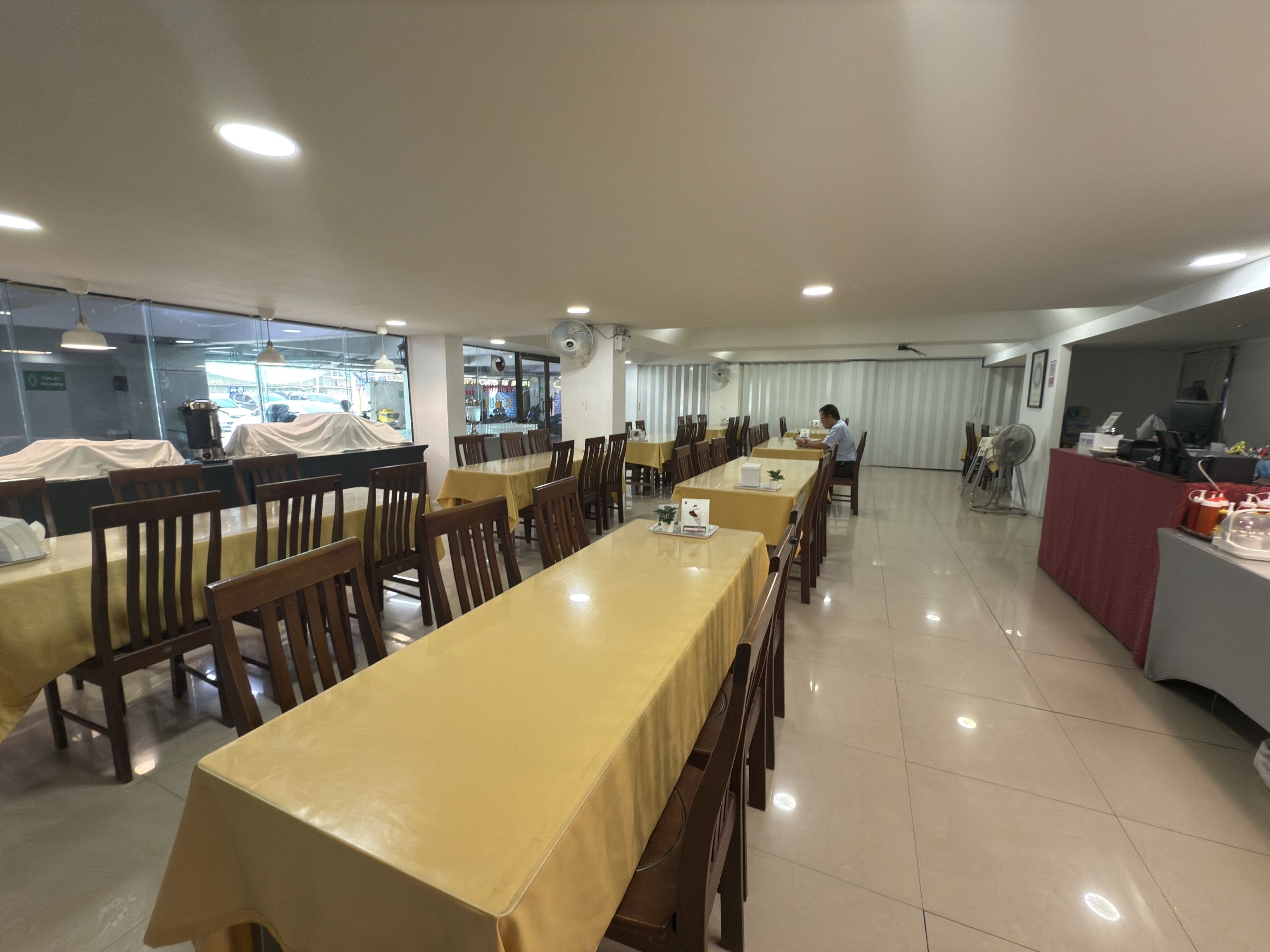 Un grand espace dédié à la salle à manger, avec de longues tables et des chaises, un coin cuisine situé à droite, et une vue sur la rue grâce à de grandes fenêtres.