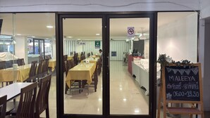 A restaurant with tables and chairs, a chalkboard menu, and a sign indicating operating hours. - Regent Ngamwongwan (Nonthaburi)