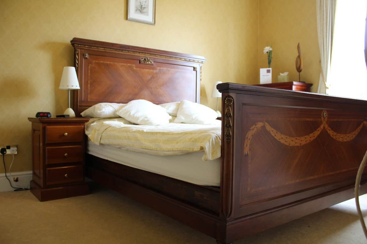 A wooden four-poster bed with a nightstand and lamp, a wall clock, and a framed picture above the bed.