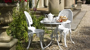 A white metal table and chairs set in a garden with a stone planter and flowers.
