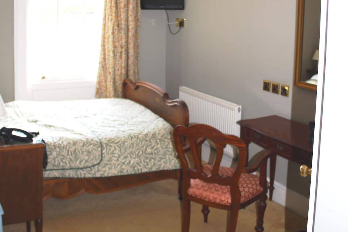 A bedroom with a bed, a wooden chair, a desk, a radiator, and a television on the wall.