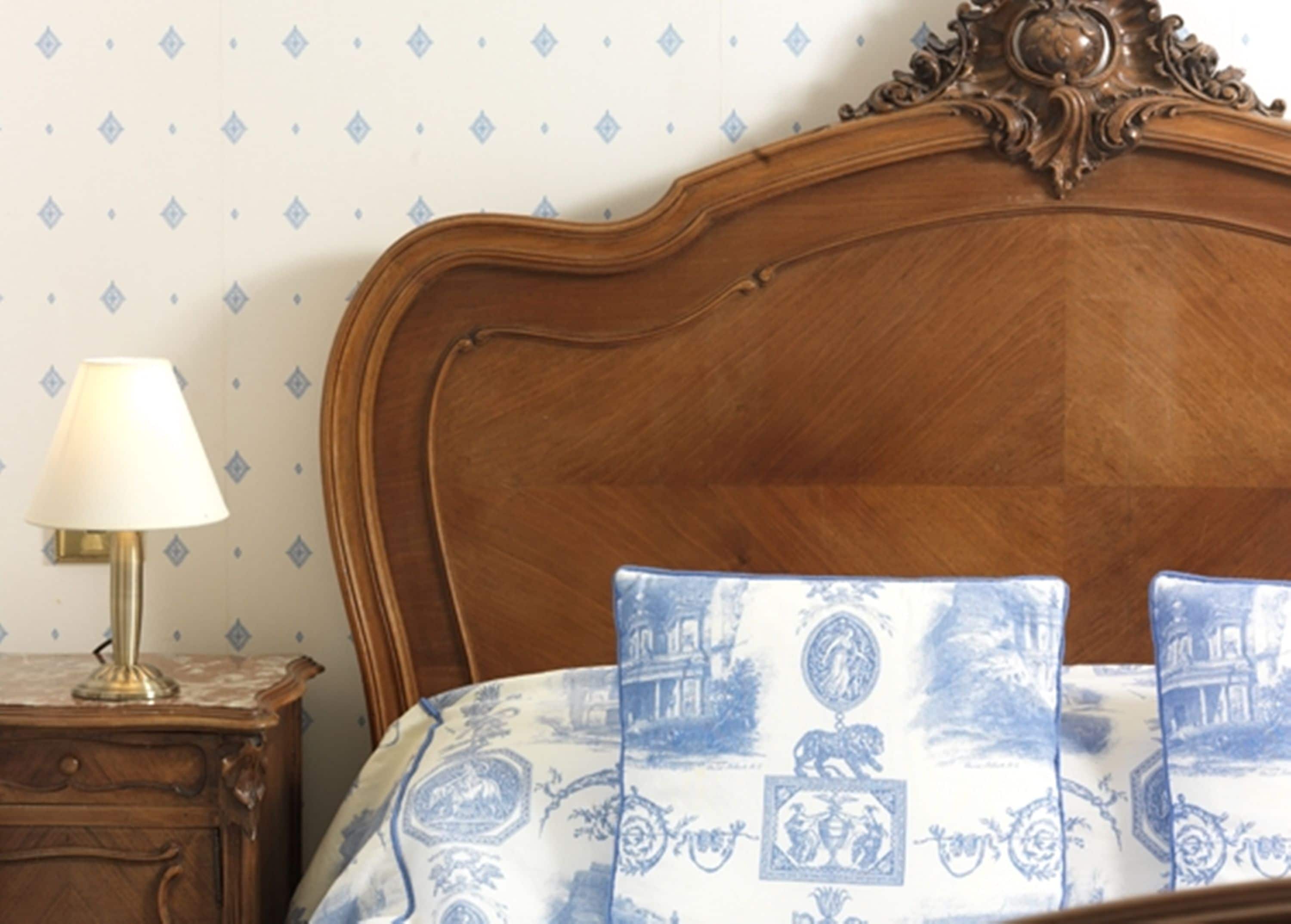Une tête de lit en bois ornée de sculptures complexes, une table de chevet avec une lampe et un lit recouvert d’une couvre-lit à motifs bleus et blancs.