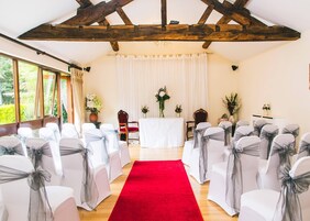 Una habitación con sillas blancas adornadas con cintas grises, una alfombra roja y una mesa blanca con arreglos florales.