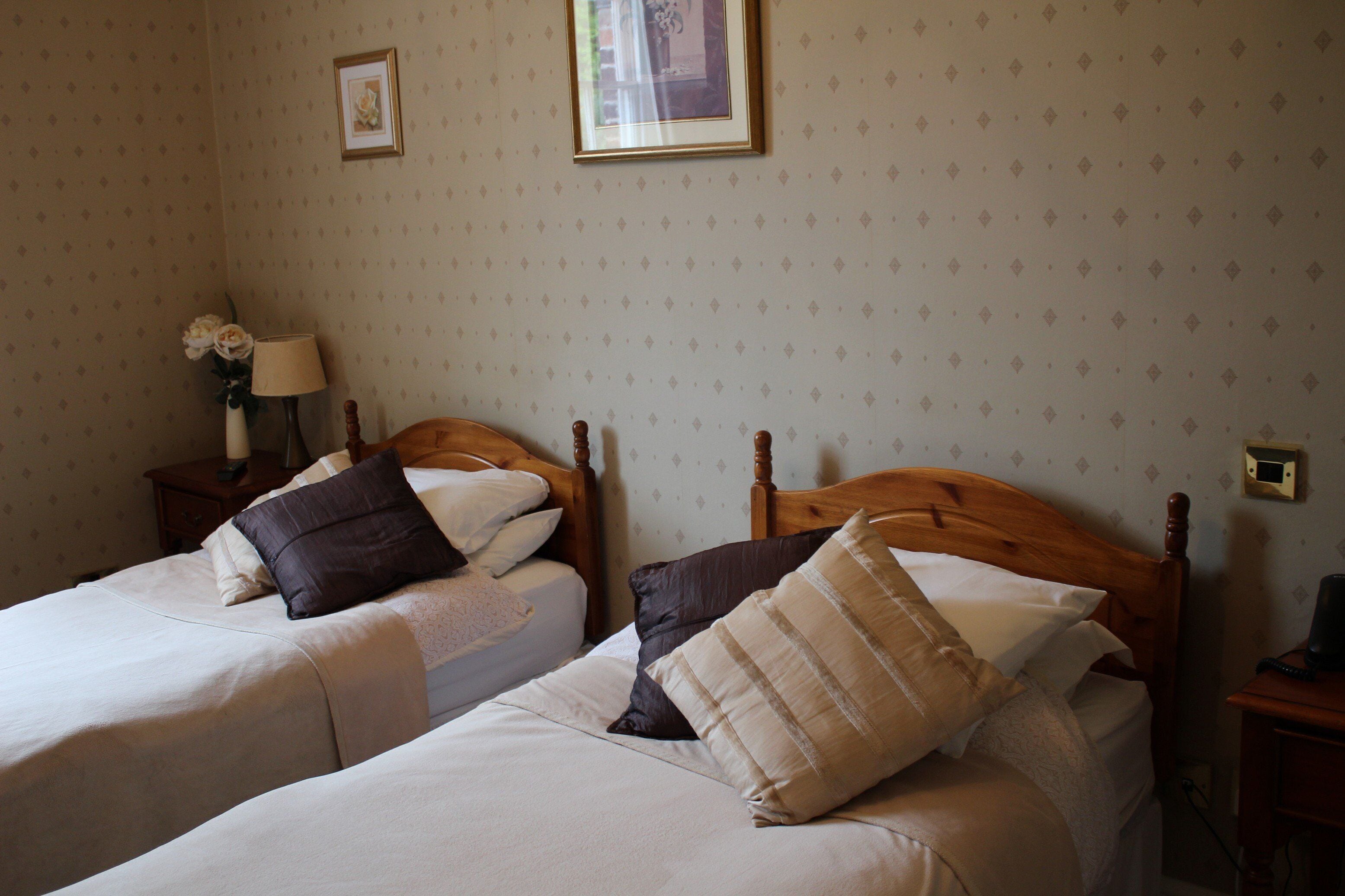 Habitación de hotel con dos camas, cada una con cabecero de madera, ropa de cama blanca y varios almohadones. Se observa una mesita de noche con lámpara y un jarrón con flores a la izquierda.