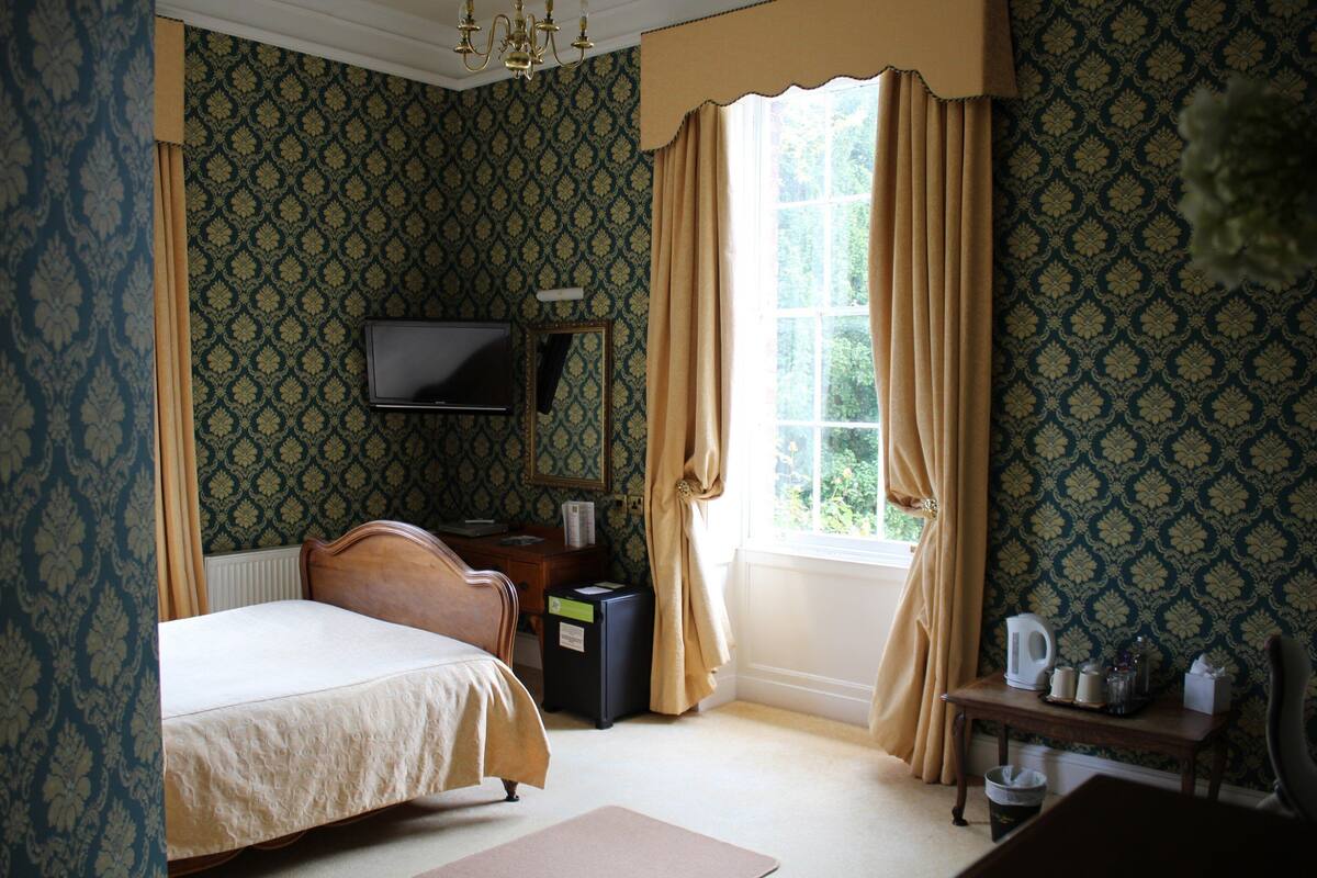 A bedroom with a bed, a dresser, a television mounted on the wall, a window with curtains, and a small table with items on it.