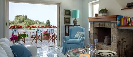 A living room with a sofa, armchair, and coffee table. There is a fireplace, books, and a view of a garden through a glass door.