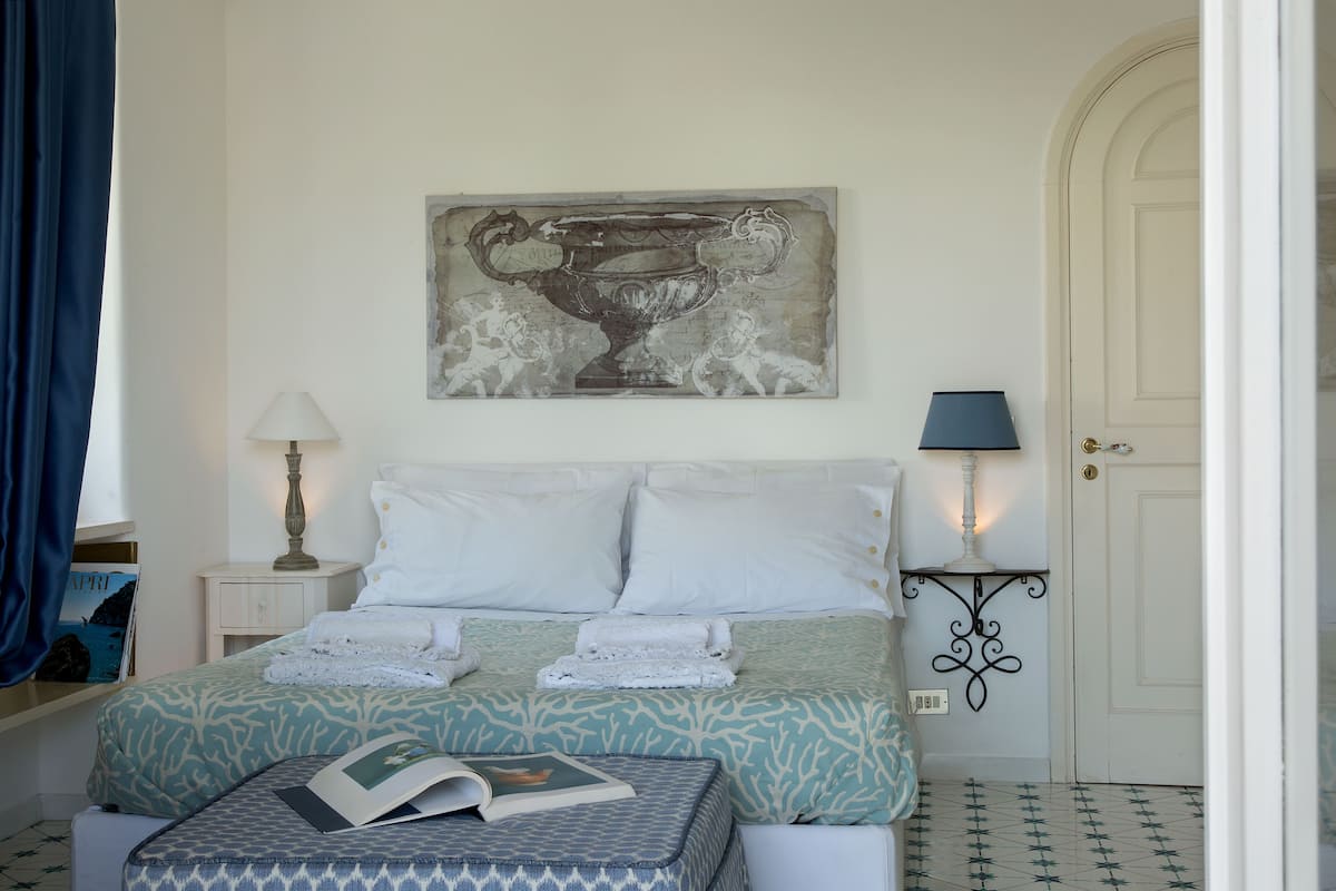 Een keurig opgemaakte slaapkamer met een bed, een nachtkastje met een lamp, een aan de muur hangend kunstwerk en een deur.
