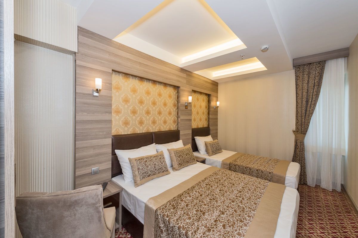 A hotel room with two beds, a desk, and a chair. The room has a modern design with patterned wallpaper and a ceiling light fixture.