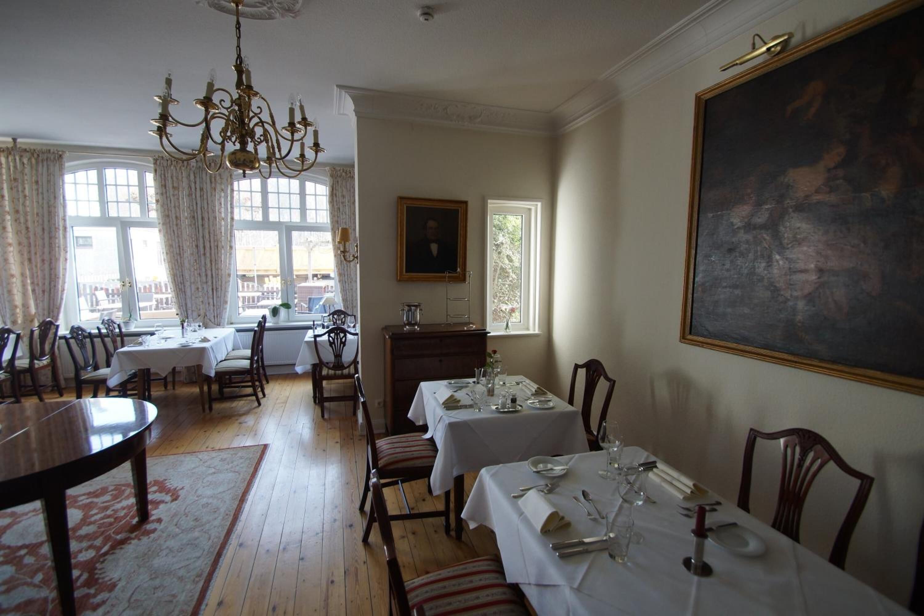 Une salle à manger agrémentée d’un lustre, avec des tables dressées pour le repas et un grand tableau accroché au mur.