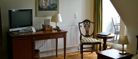 A hotel room with a wooden desk, a television, a chair, a bedside table, and a bed.