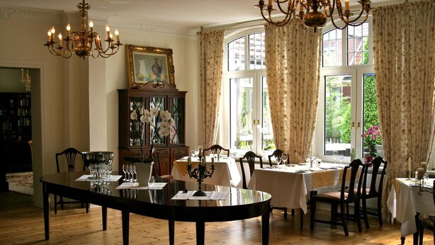 A dining area with a large round table, multiple chairs, and a chandelier. There is a window with curtains, a cabinet with glass doors, and a painting on the wall. - Landhaus Bode (Lübeck)