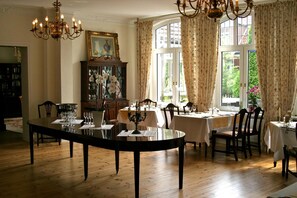 A dining area with a large round table, multiple chairs, and a chandelier. There is a window with curtains, a cabinet with glass doors, and a painting on the wall. - Landhaus Bode (Lübeck)