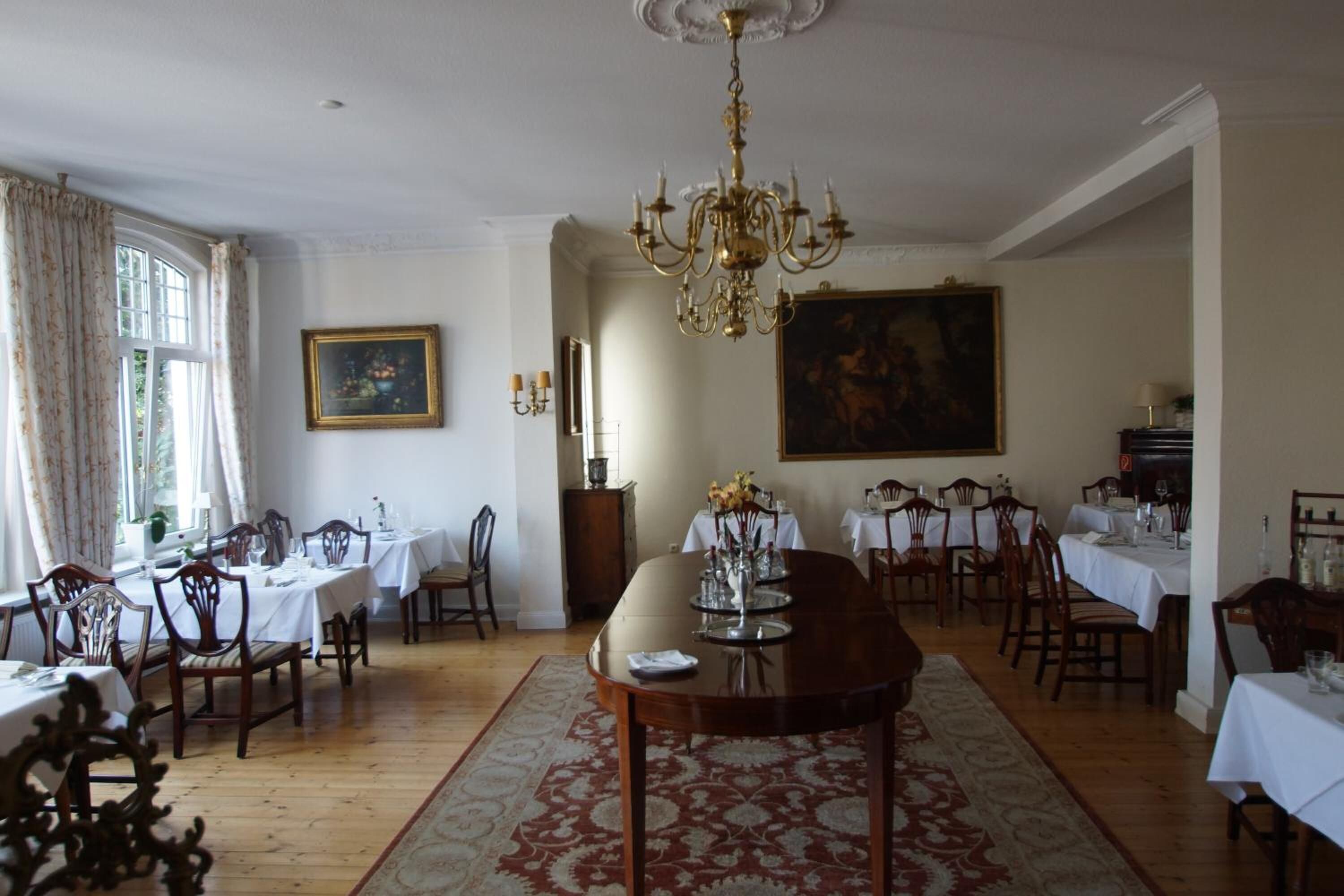 A dining room with a large wooden table, chairs, and a chandelier. There are paintings on the walls and a window with curtains.