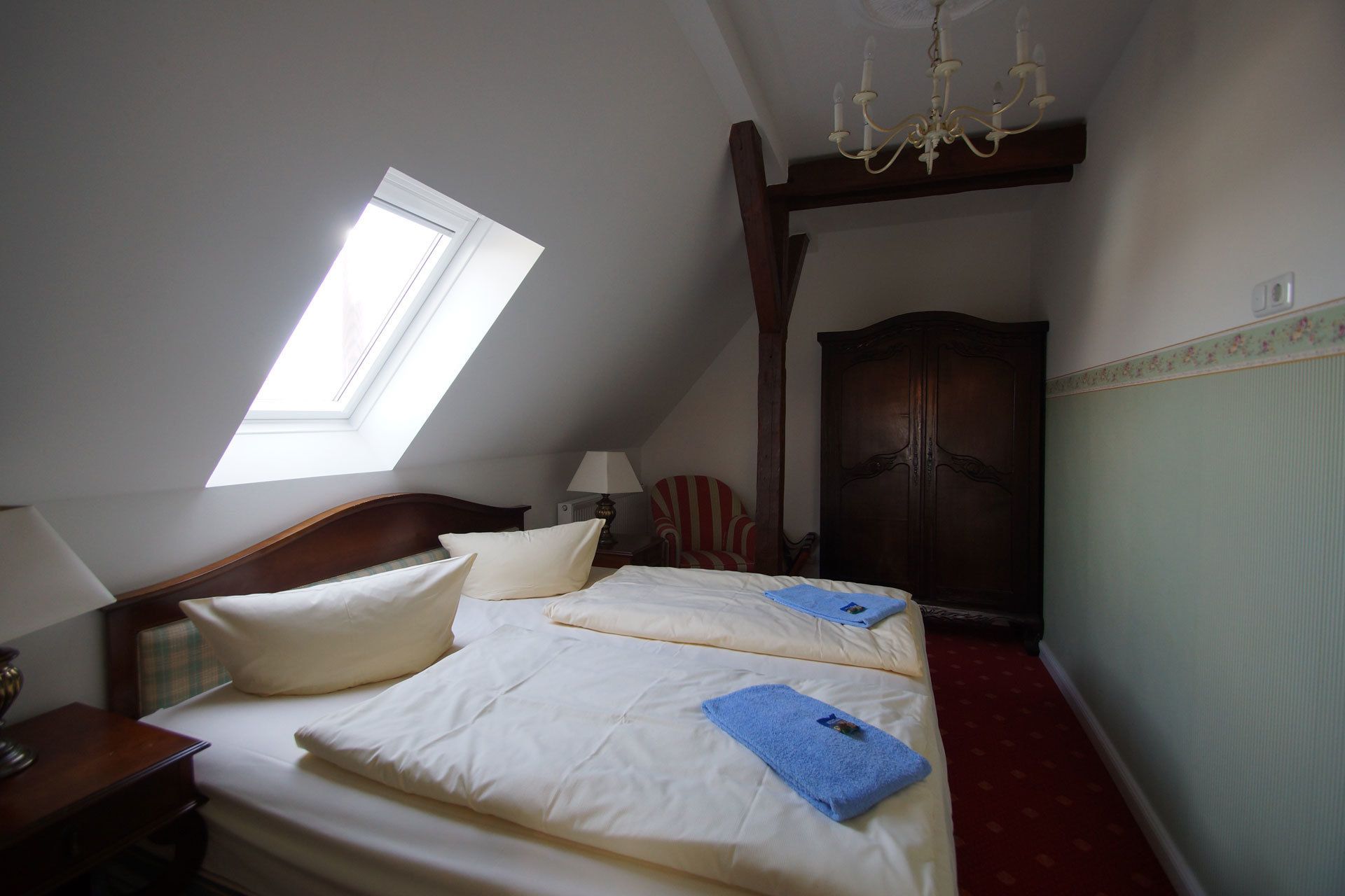 A bedroom with a skylight, a chandelier, a wooden wardrobe, and a bed with pillows and a blue towel.