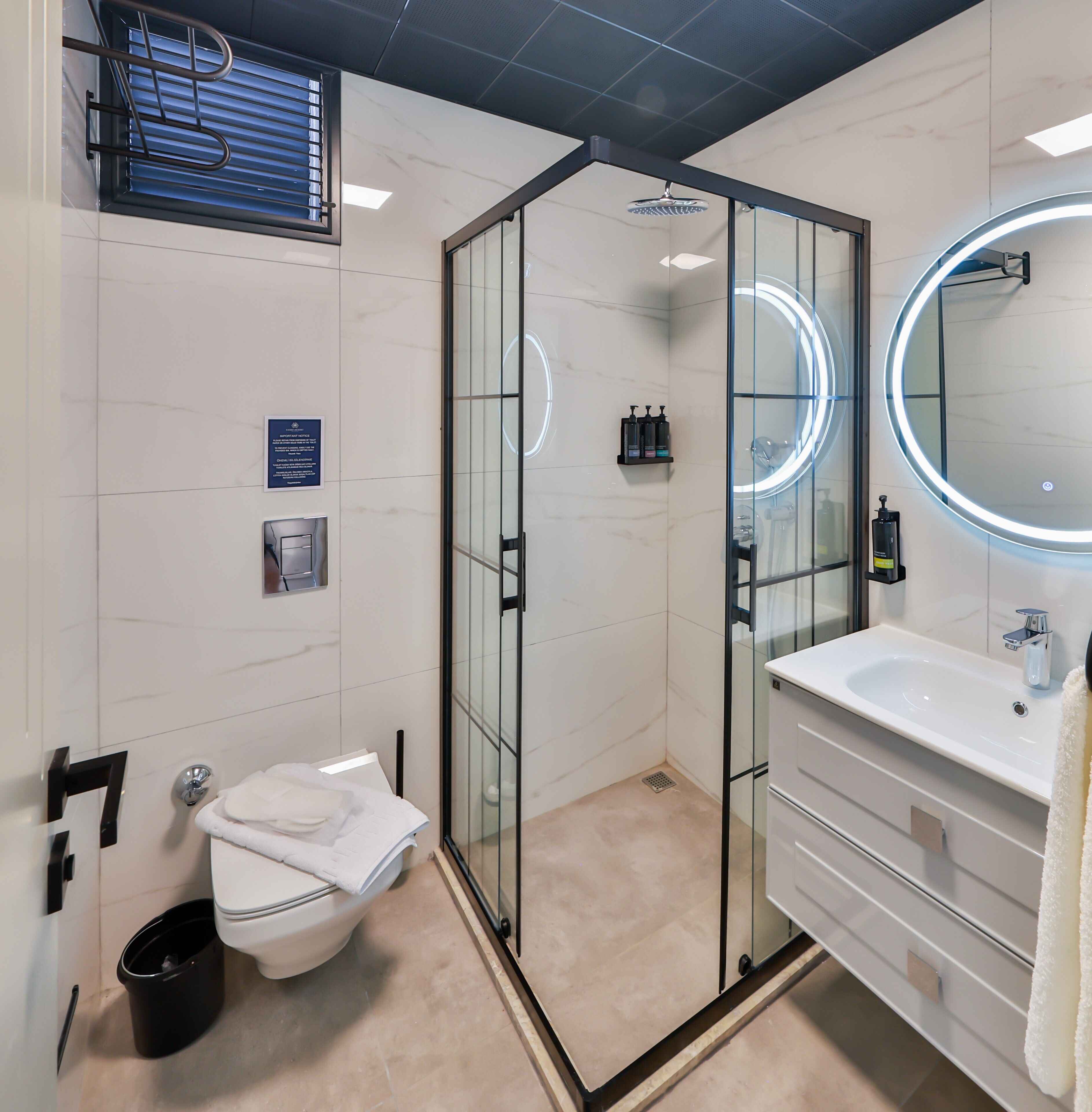 A modern bathroom with a glass-enclosed shower, a white sink, and a mirror with a circular light.