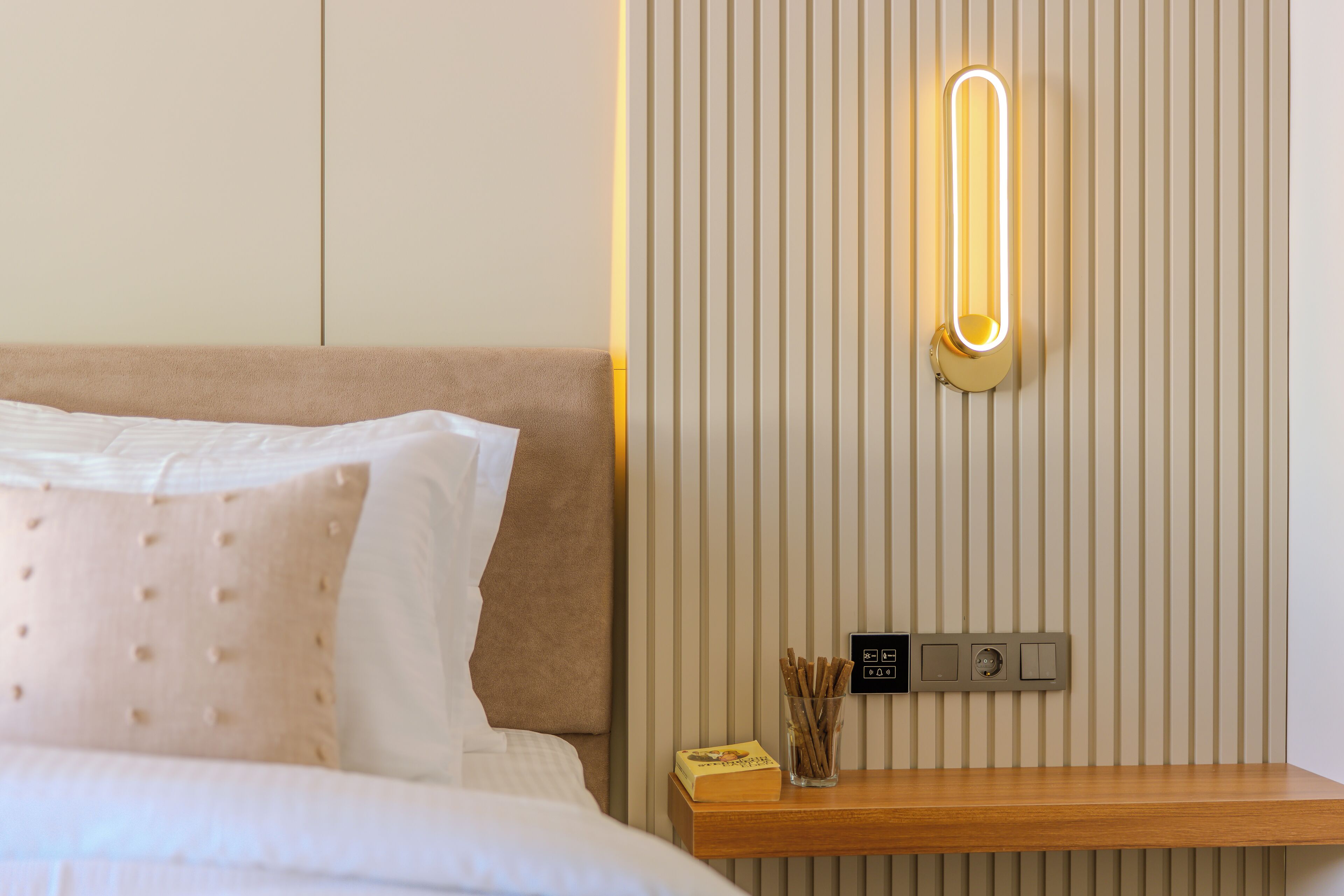 Een net opgemaakt bed met hoofdbord, nachtkastje met lamp en een wandlamp.