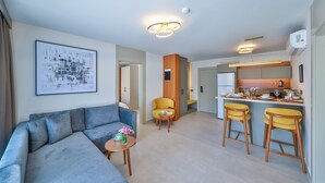 A modern living room with a sofa, a small table with a cup and flowers, a yellow armchair, and a kitchen area with a bar.