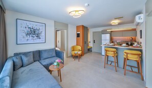 Uma sala de estar moderna com um sofá, uma mesinha com uma chávena e flores, um poltrona amarela e uma área de cozinha com um balcão.