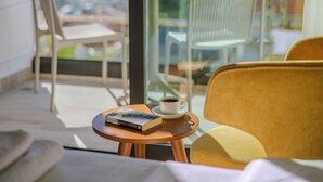 A cozy seating area with a yellow chair, a wooden table with a book and a cup of coffee, and a view through a glass door.
