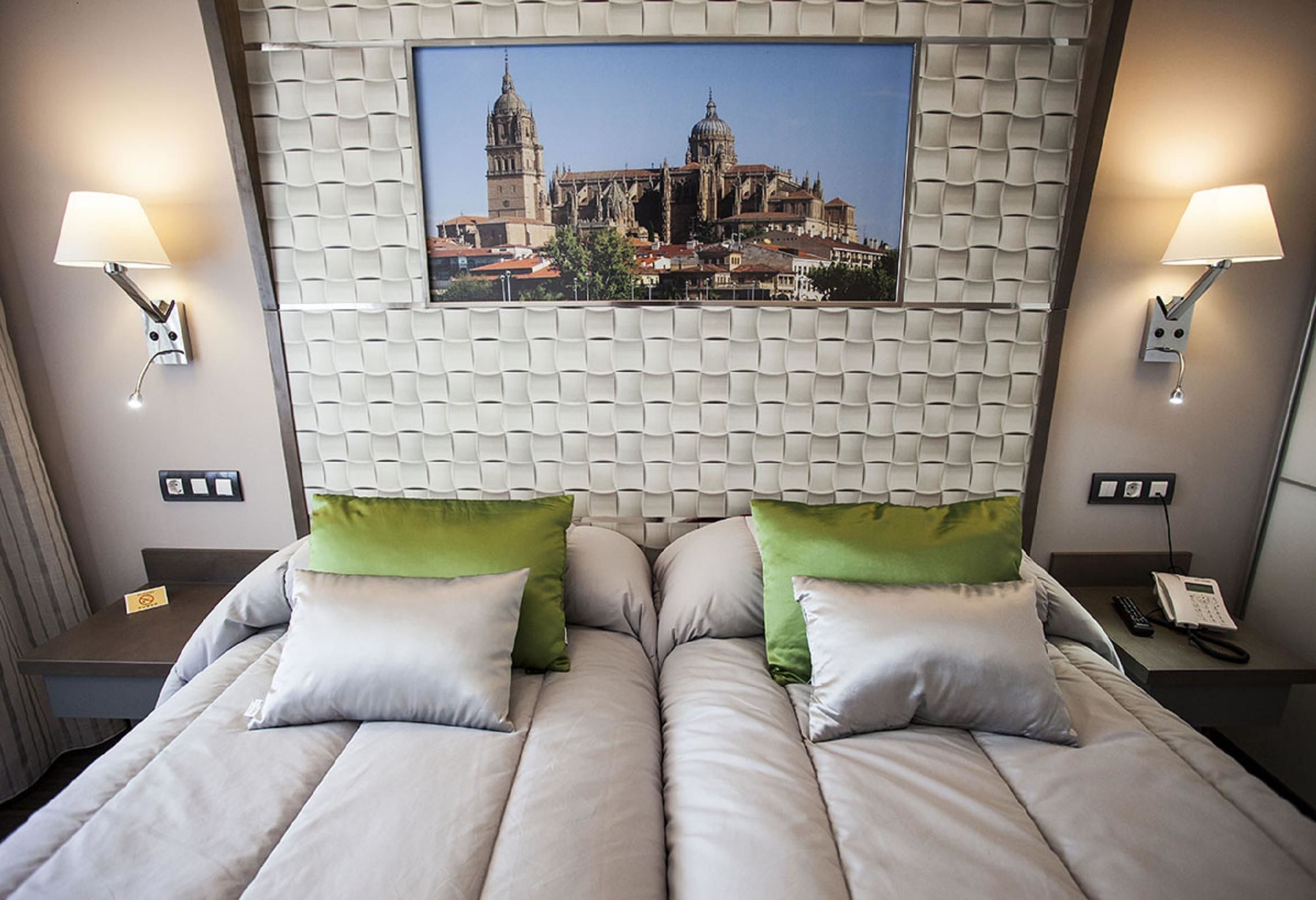 A hotel room with a large bed, two bedside tables, a wall-mounted picture of a cathedral, and two wall-mounted lamps.