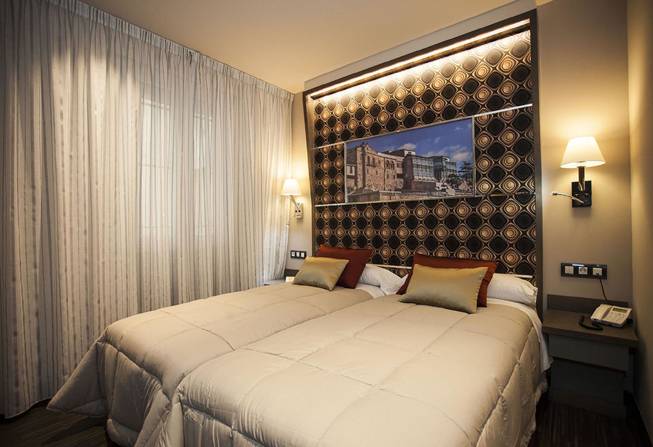 Habitación de hotel con una cama grande, un cabecero estampado, dos lámparas de noche y vistas a un paisaje urbano a través de una ventana.