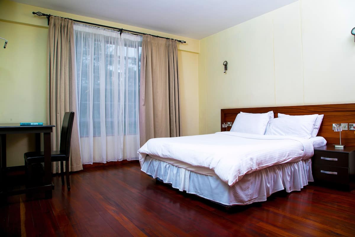 A hotel room with a wooden floor, a bed with white bedding, a desk with a chair, and a window with curtains.