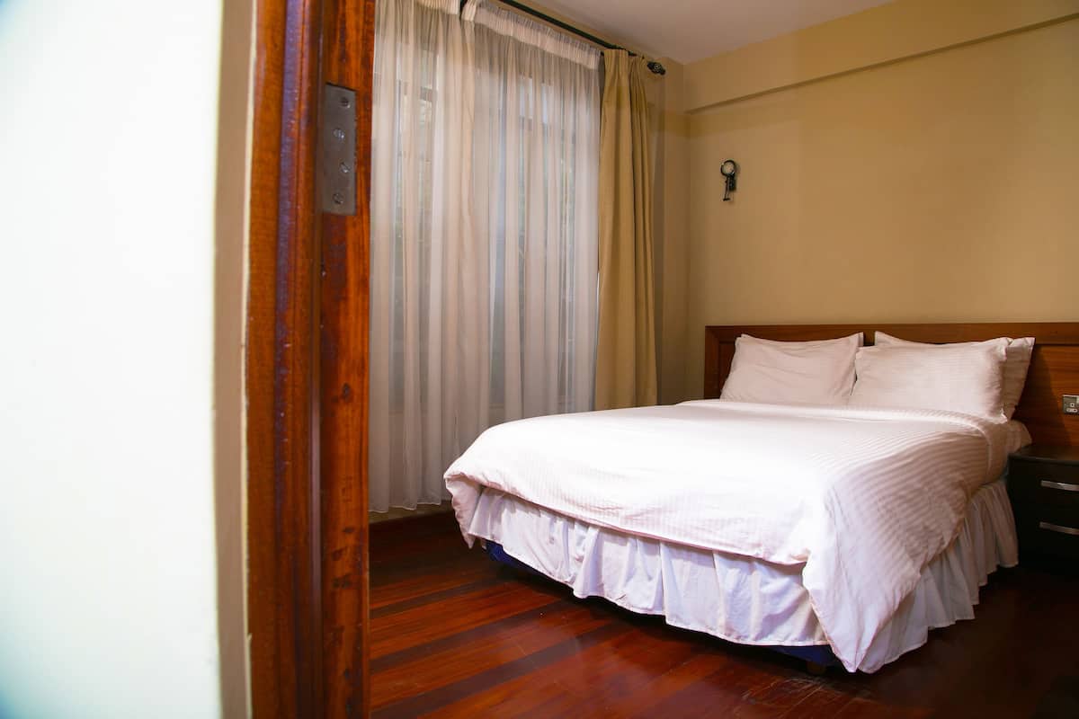 A neatly made bed with a wooden headboard, a wooden floor, and a window with sheer curtains.