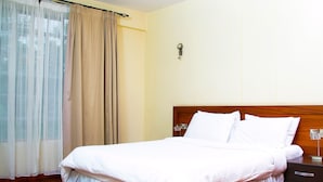A hotel room with a wooden floor, a bed with white bedding, a nightstand, and a window with sheer curtains.