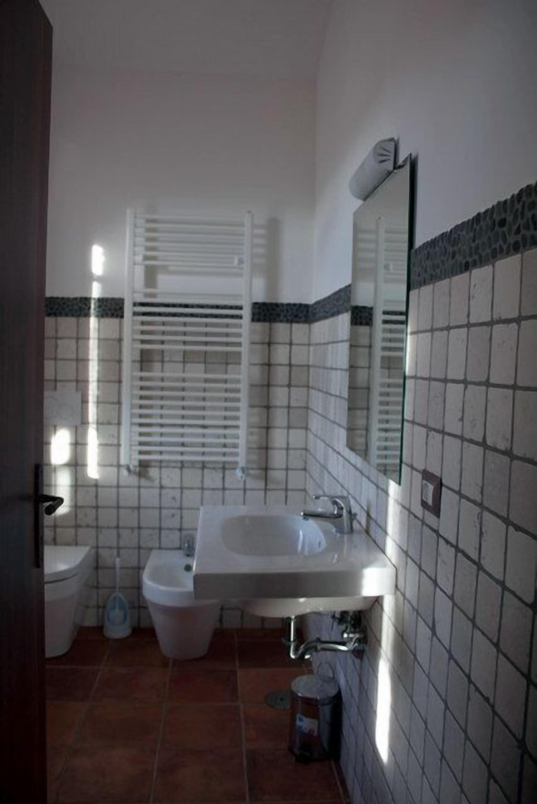 Un baño con lavamanos, bidé e inodoro. Se observa una ventana con persianas, un espejo y una pared con azulejos.