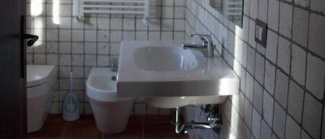 Un baño con lavamanos, bidé e inodoro. Se observa una ventana con persianas, un espejo y una pared con azulejos.